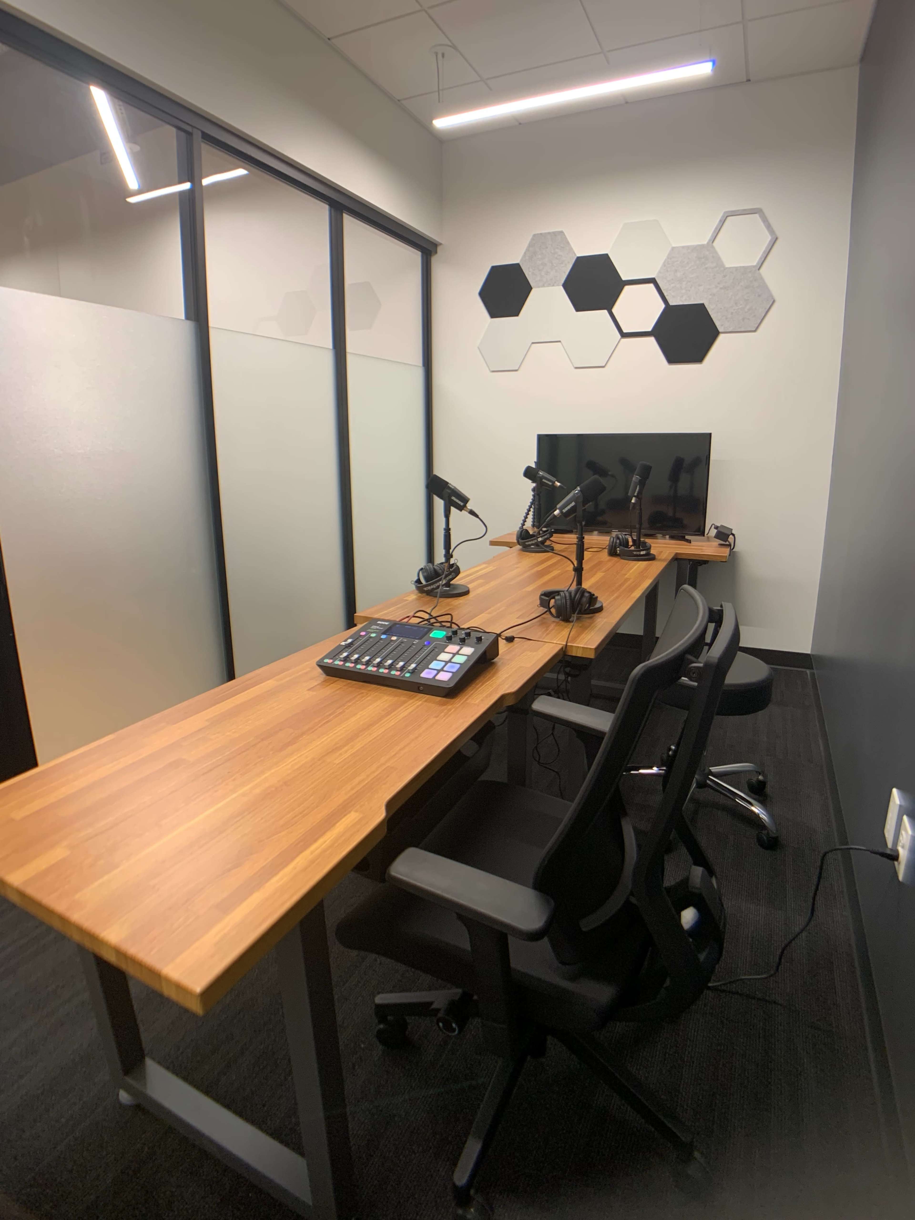 A small, modern workspace with a wooden tabletop, two chairs, a computer monitor, and multiple microphones arranged for recording or broadcasting.