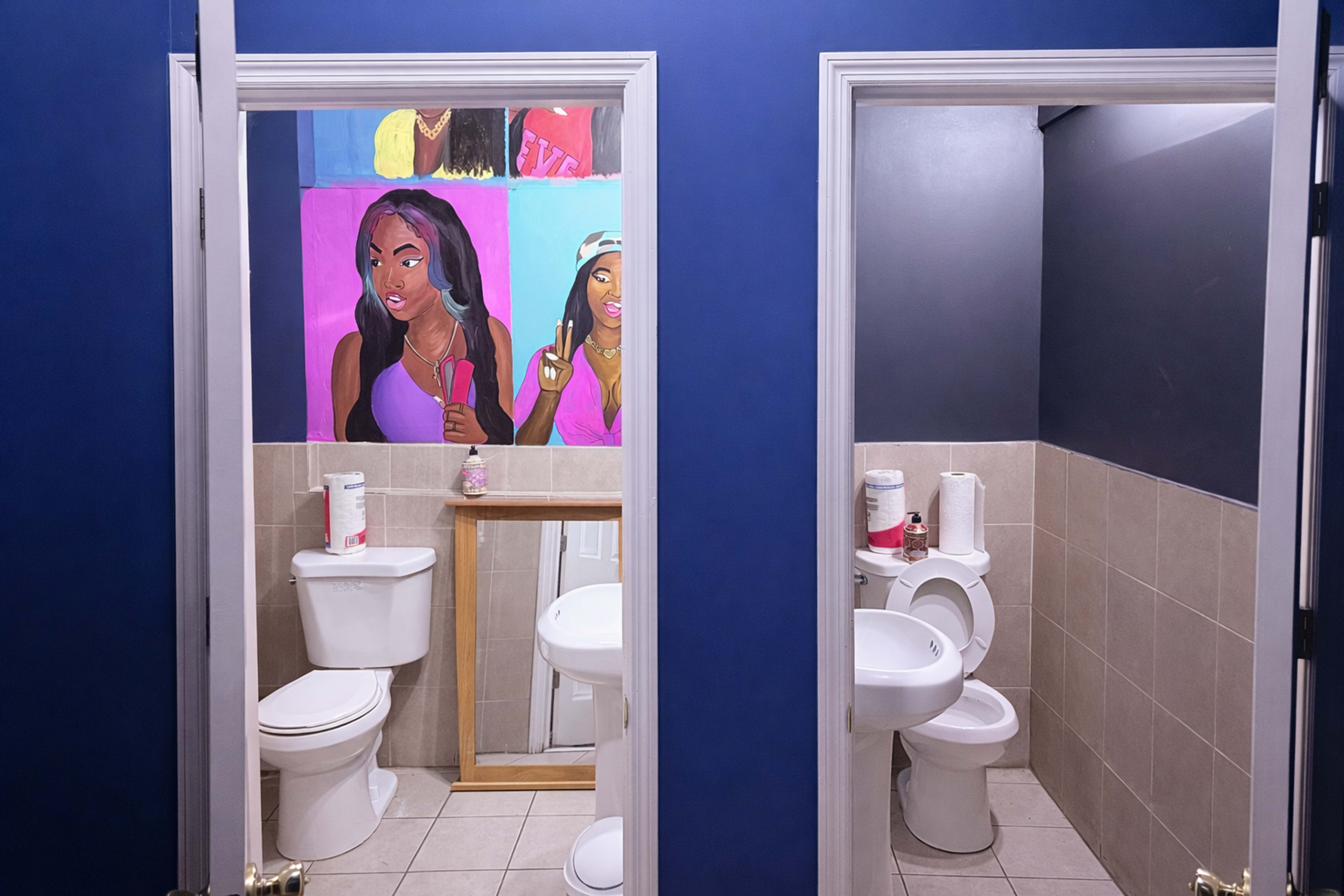 The image shows two restrooms side by side with a mural of two women on the wall between them.