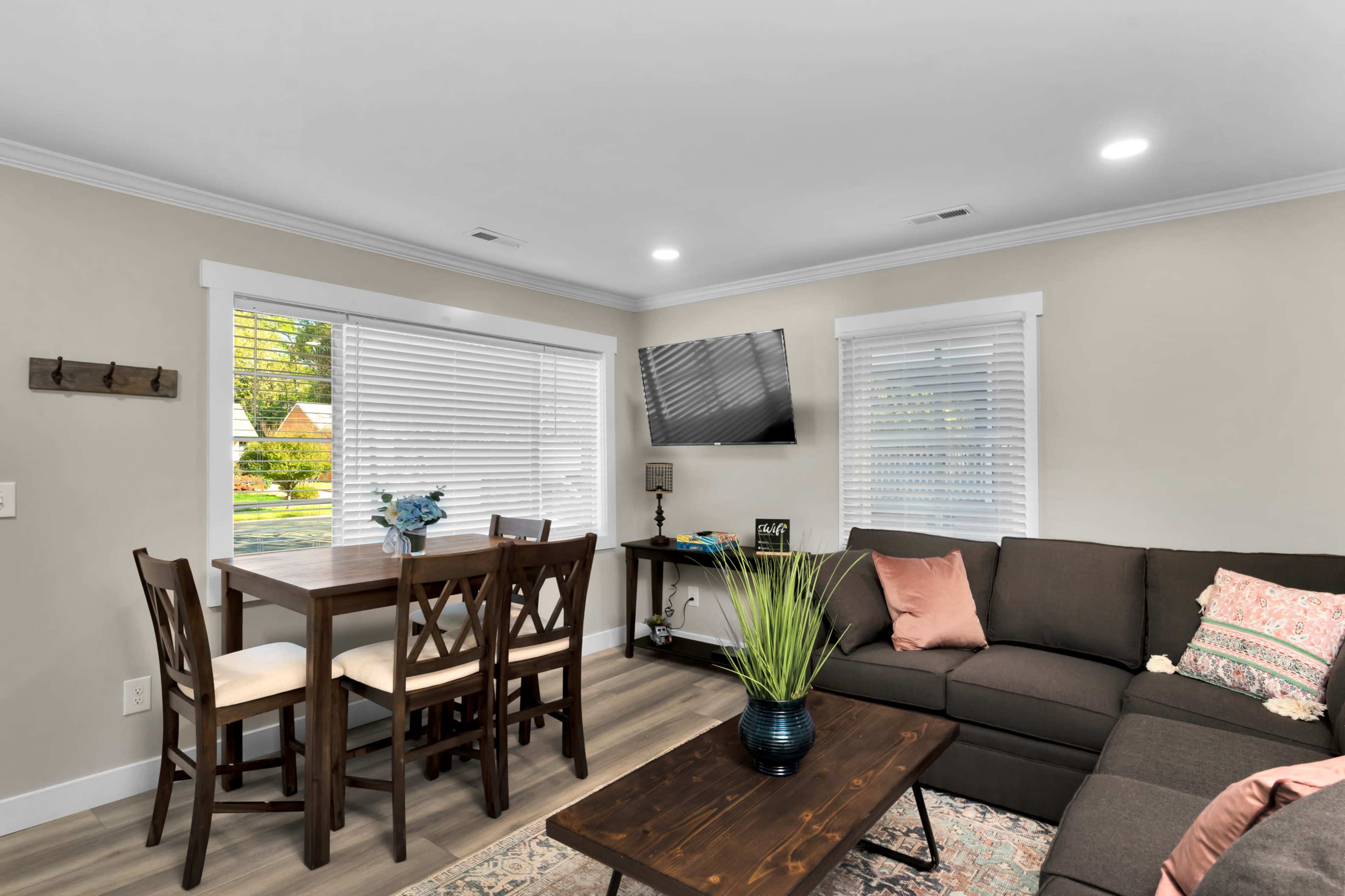A modern living room with a brown sectional sofa, a wooden coffee table, and a small dining table with chairs near a window.