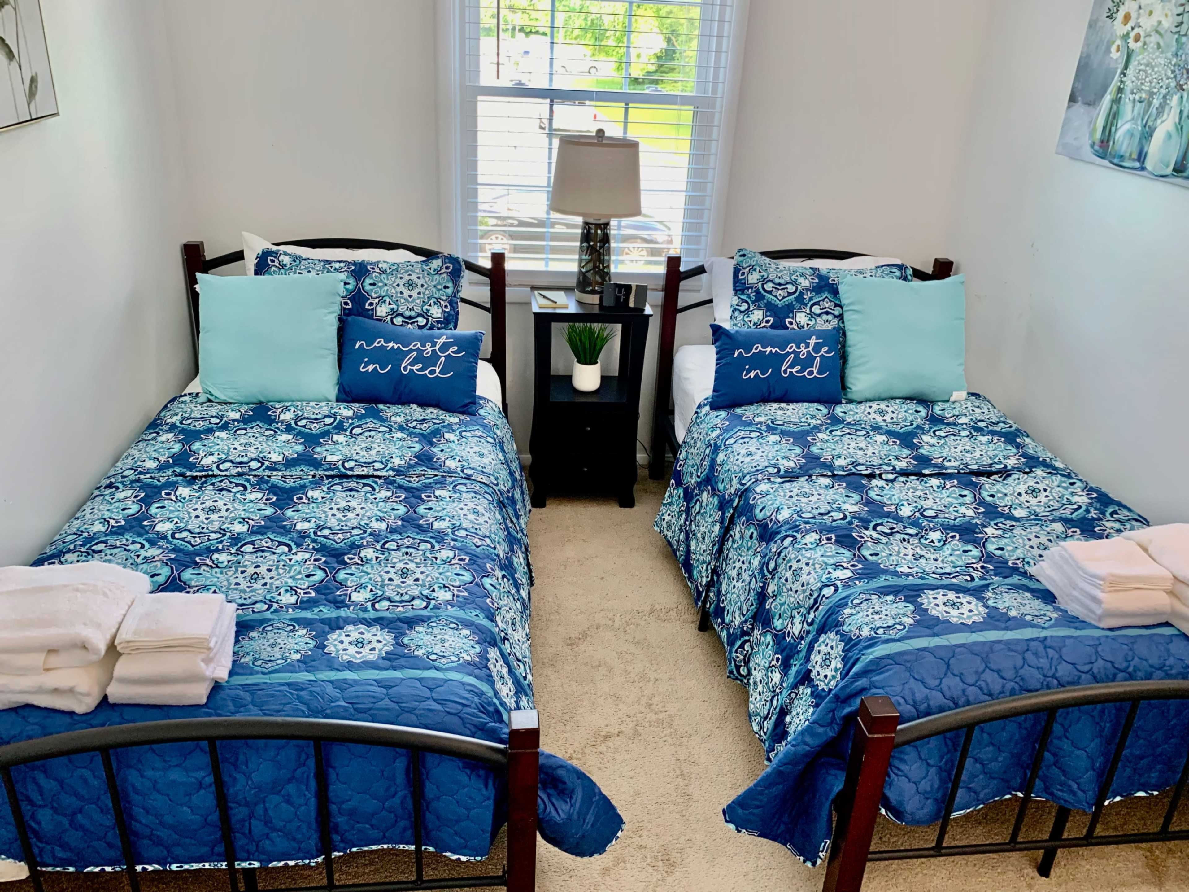 A bright bedroom featuring two twin beds with blue patterned bedding, flanked by a bedside table with a lamp and a decorative plant.