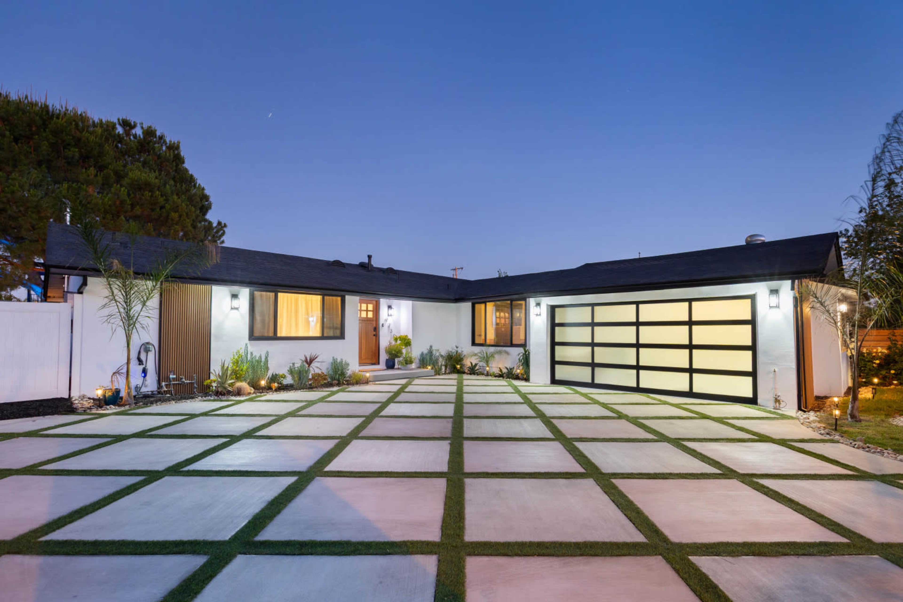 The image shows a modern single-story house with a sleek design, featuring a square driveway and landscaped patches of grass.