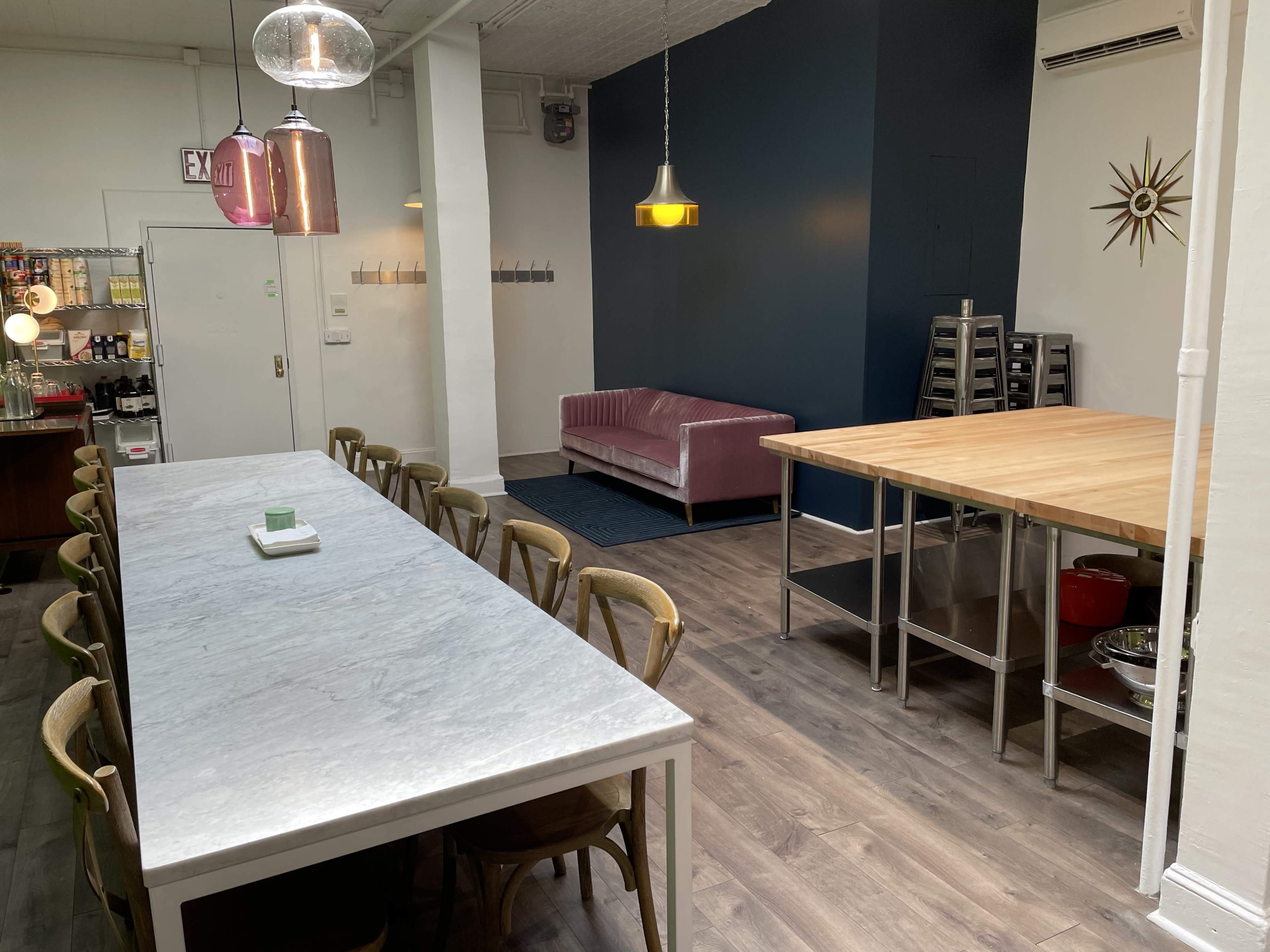 A modern kitchen space featuring a long marble dining table, a pink sofa against a blue wall, and a wooden worktable.