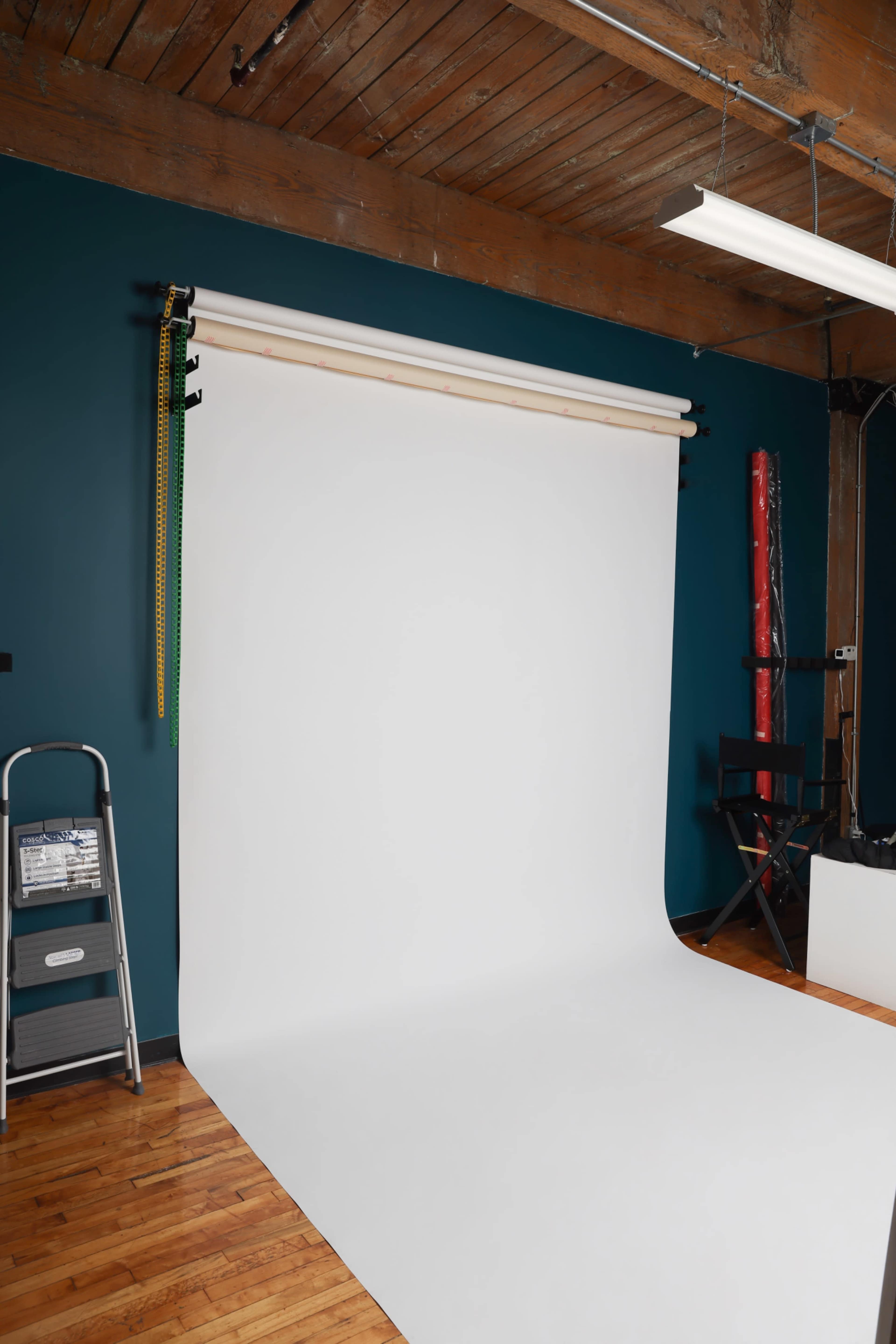 The image shows a photography studio with a white backdrop, a metal step ladder, and wooden beams on the ceiling.