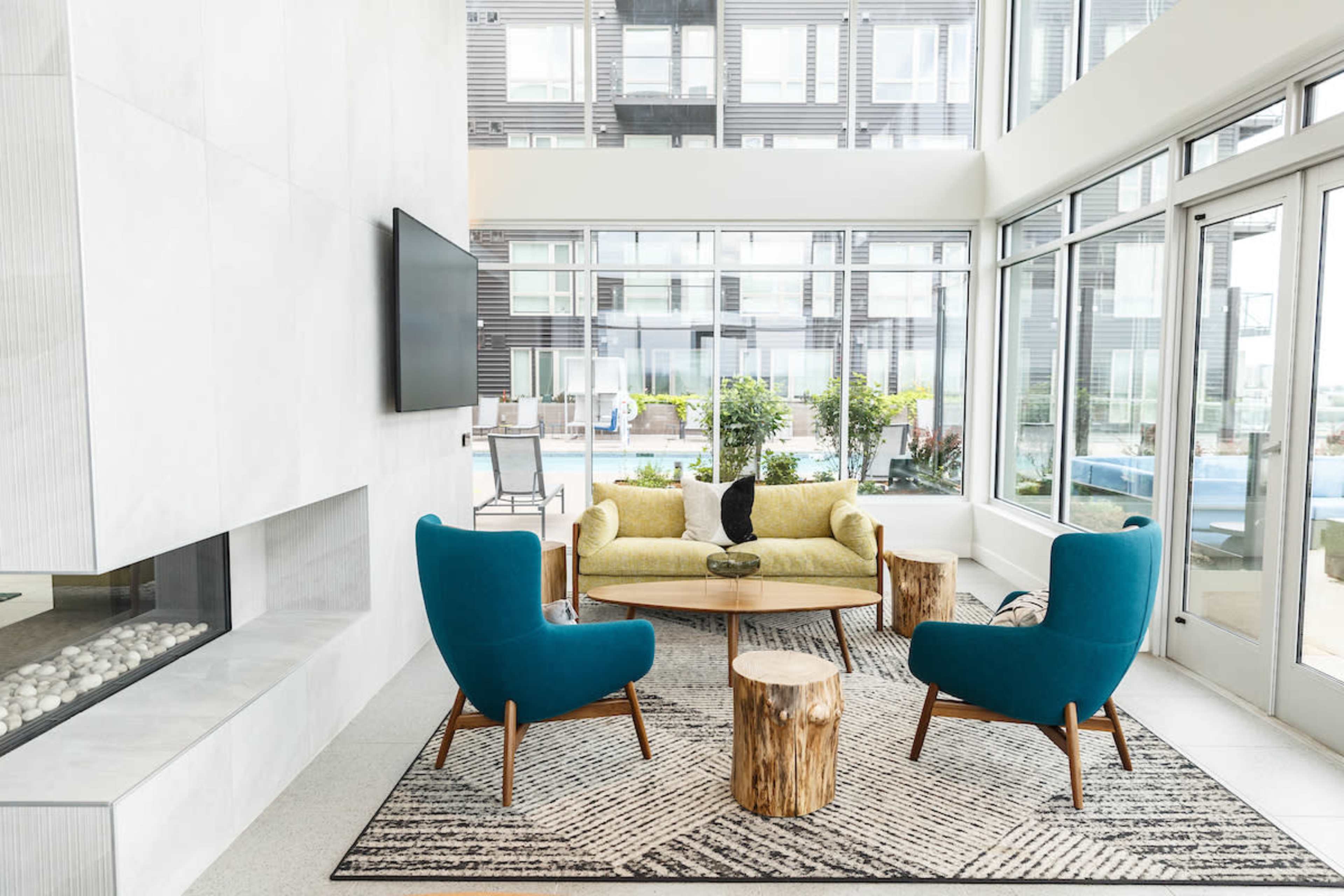 A modern living area features a yellow sofa, turquoise armchairs, a wooden coffee table, and large windows overlooking an outdoor pool area.
