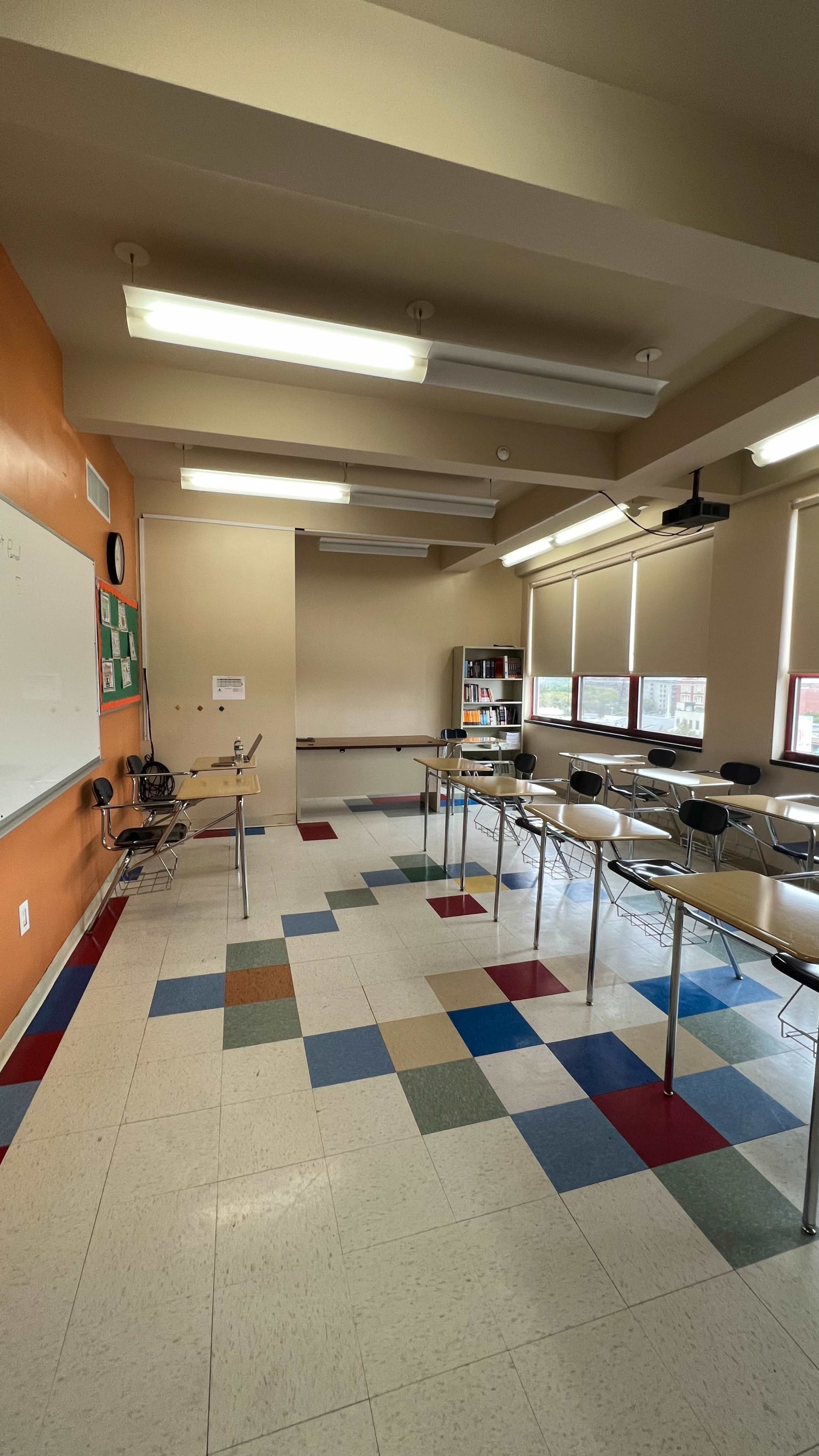 The image shows a classroom with colorful floor tiles, a whiteboard, desks with chairs, and large windows allowing natural light.