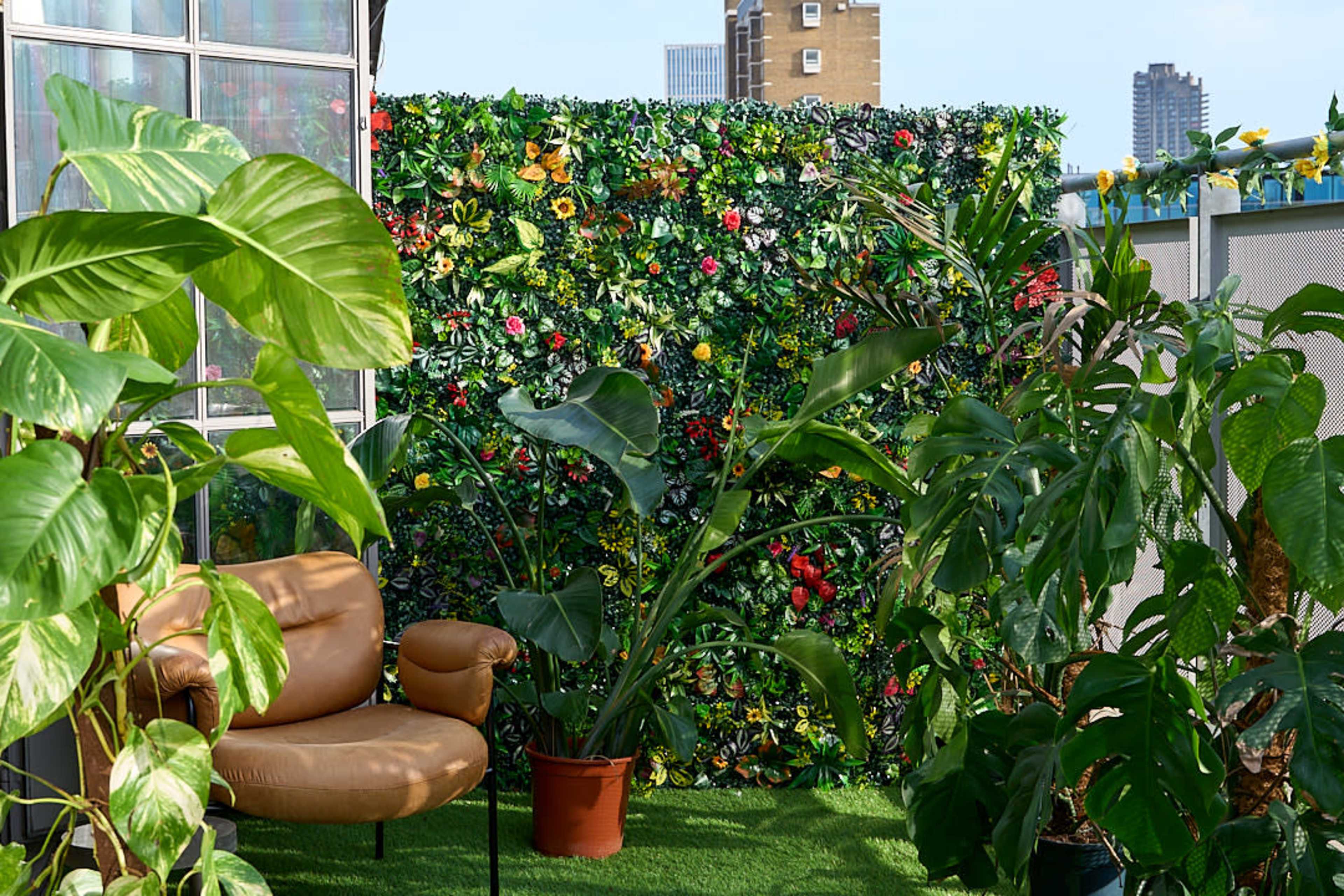 A cozy seating area features a brown leather chair in front of a vibrant green wall adorned with various plants and flowers, with additional potted plants surrounding the space.