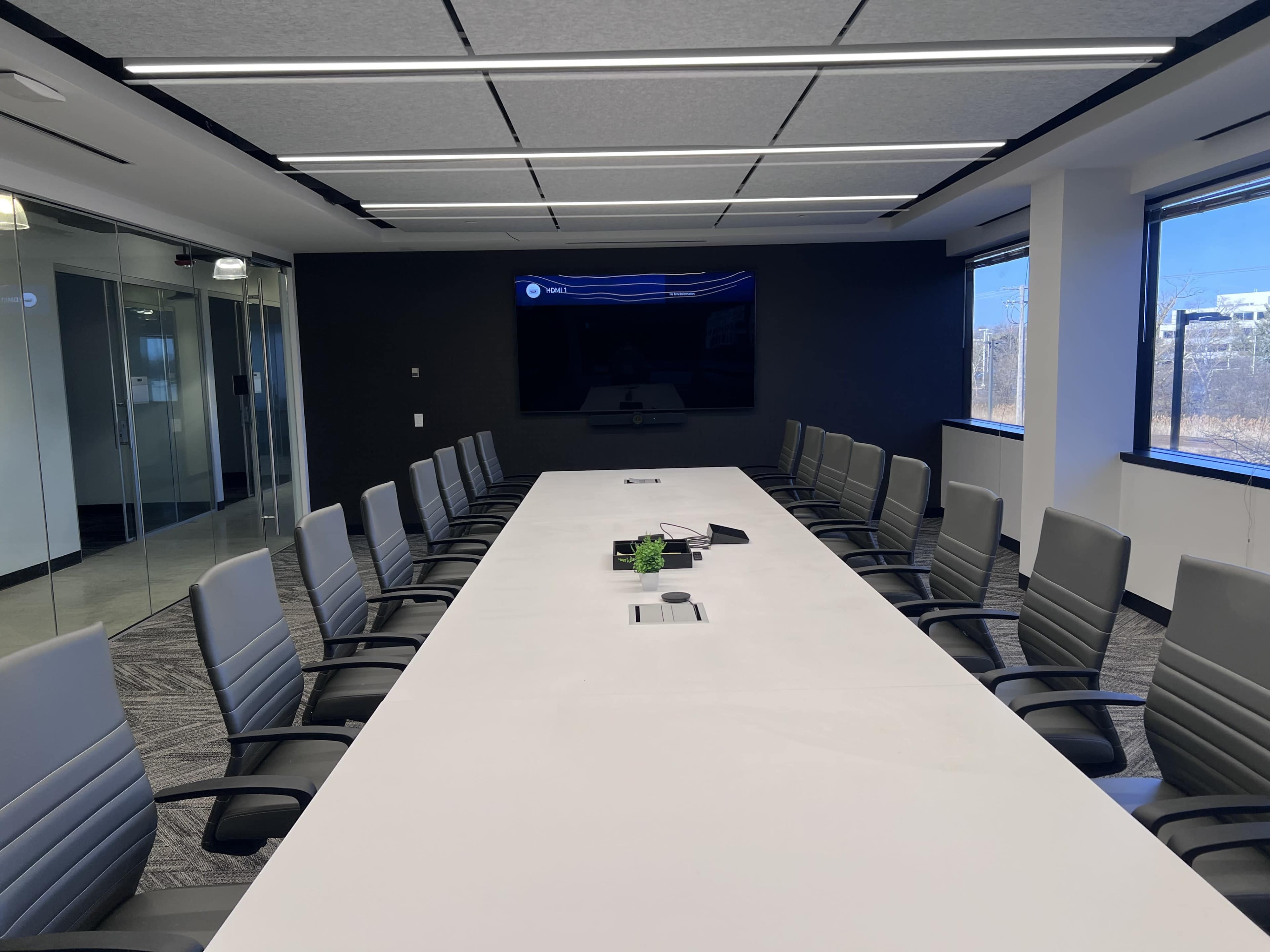 A large conference room features a long white table surrounded by gray chairs, with a television mounted on a black wall and windows allowing natural light.