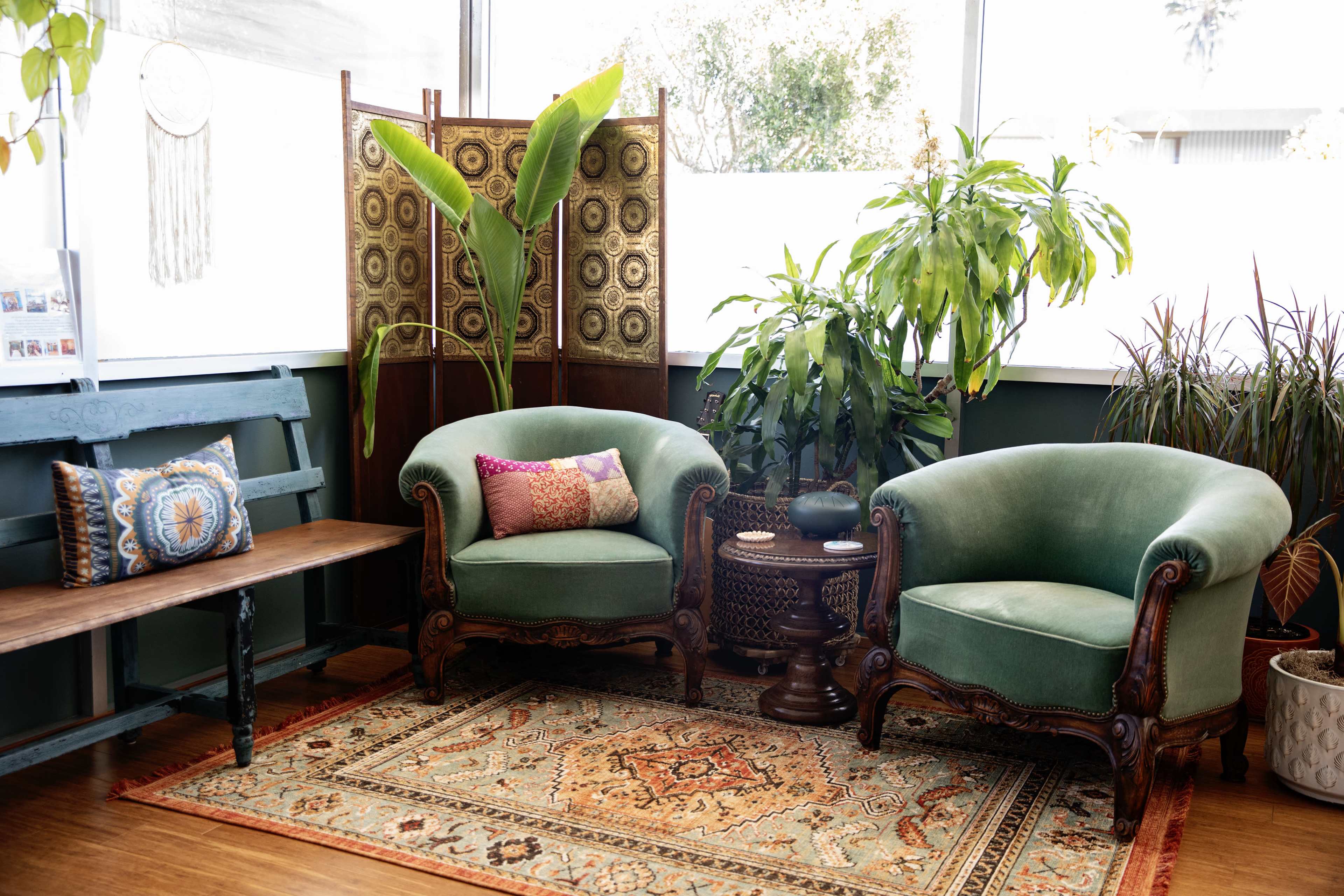 The scene features two green upholstered armchairs, a small circular wooden table, and various plants arranged in a bright room with large windows.