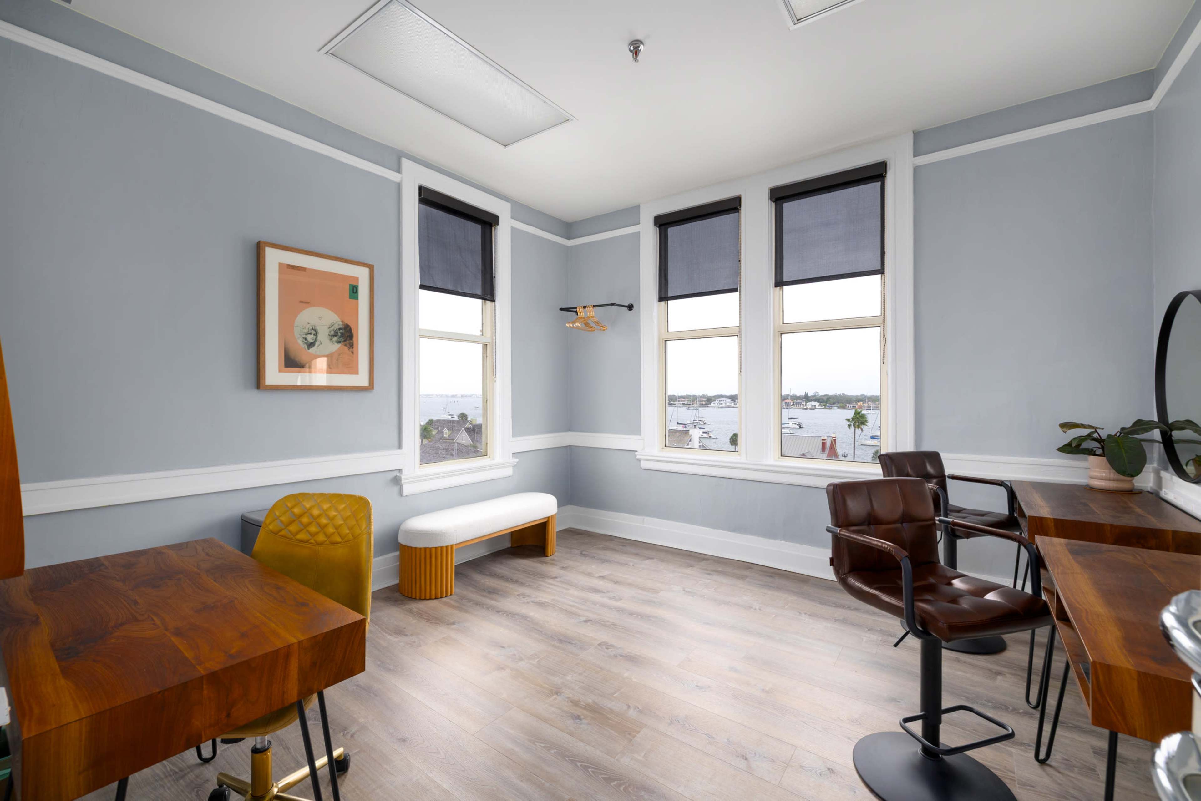 The image shows a bright, minimalistic room with two wooden desks, a bench, and large windows providing a view of the outside.