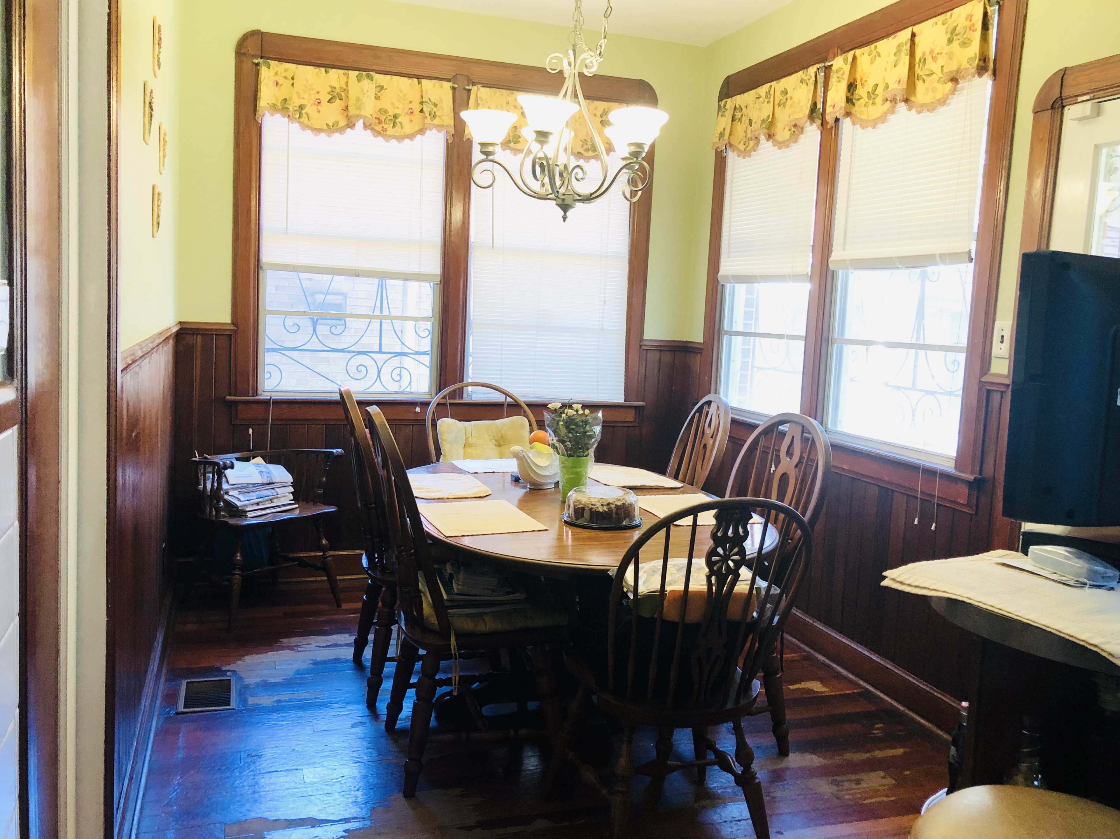 A dining room features a wooden table surrounded by chairs under a chandelier, with windows adorned with floral valances.