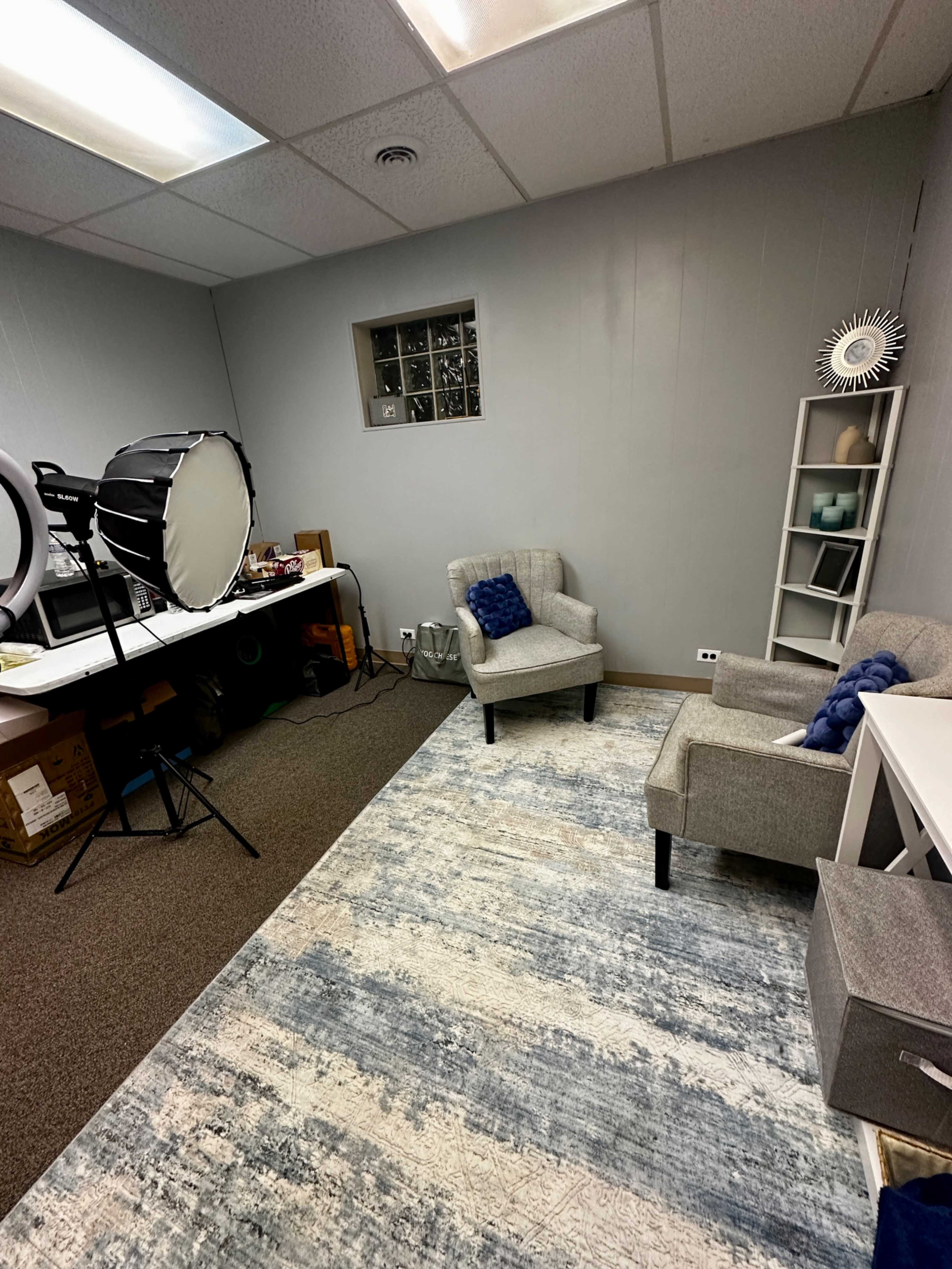 The image shows a small room with gray walls, two gray chairs, a light source on a stand, and a storage shelf with decorative items.