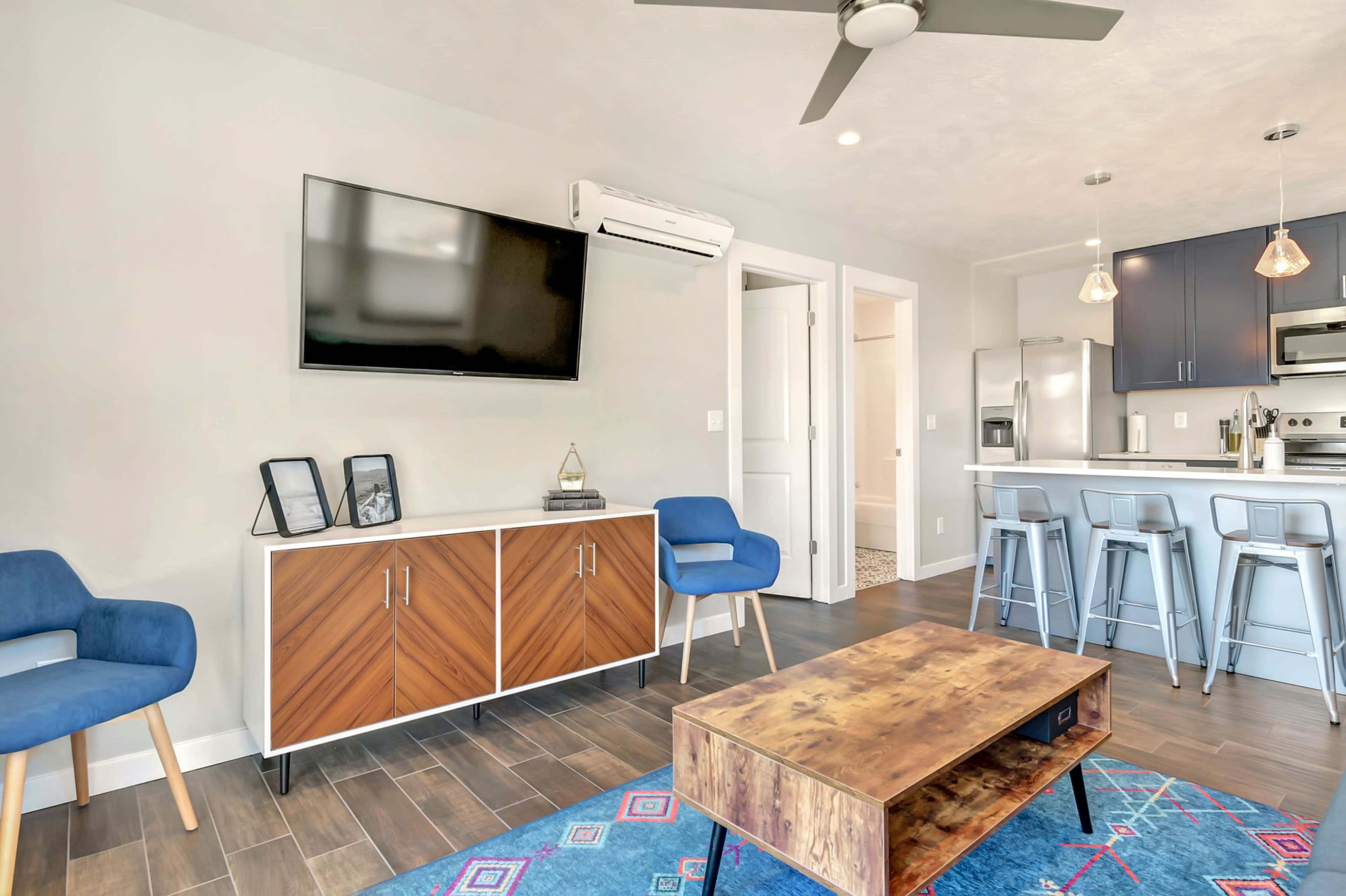 The scene depicts a modern living area featuring a wall-mounted TV, two blue chairs, a wooden coffee table, and a kitchen area with bar stools in the background.
