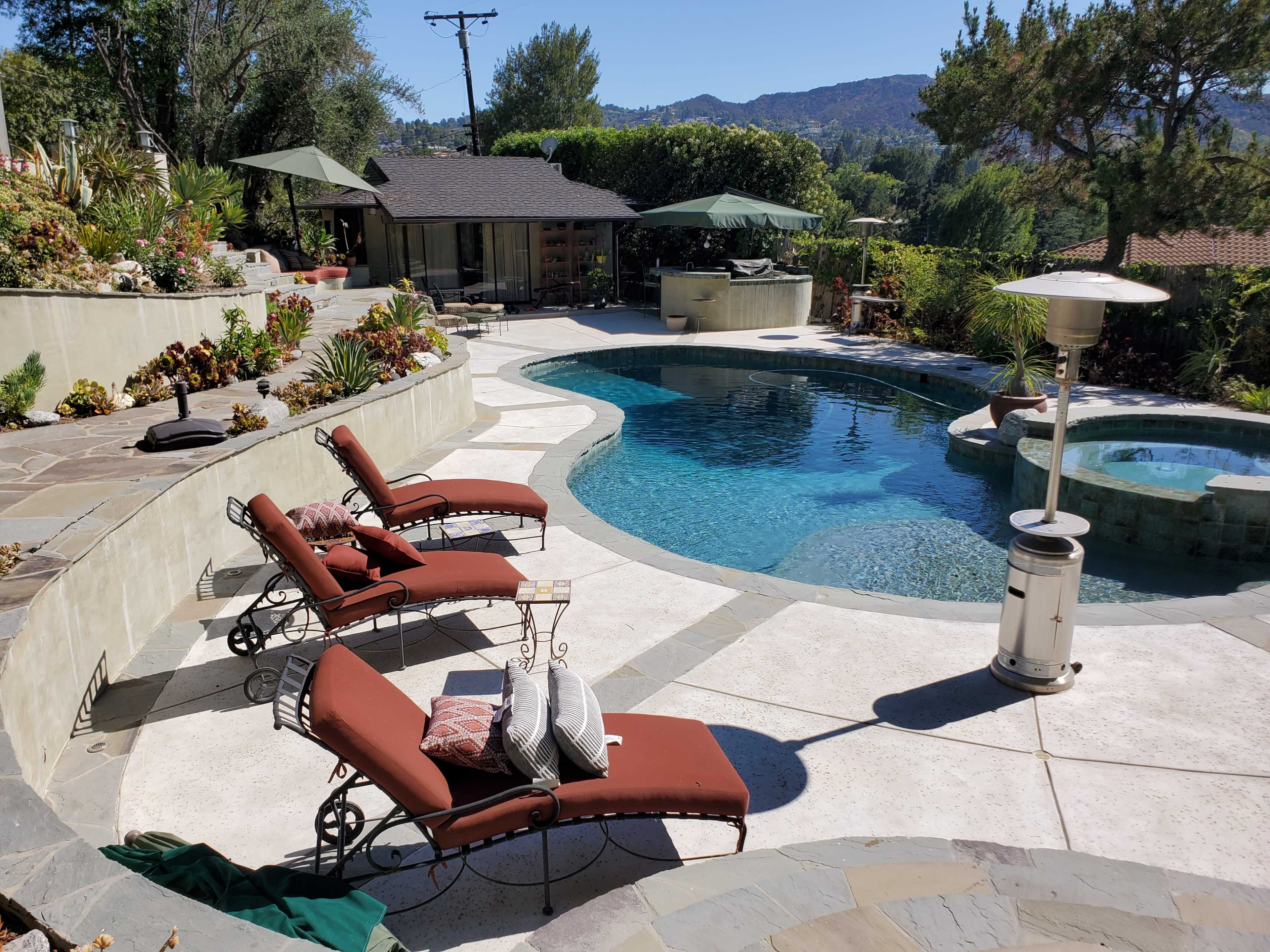 A landscaped backyard with a swimming pool, two lounge chairs, a spa, and several umbrellas surrounded by greenery and mountains in the background.