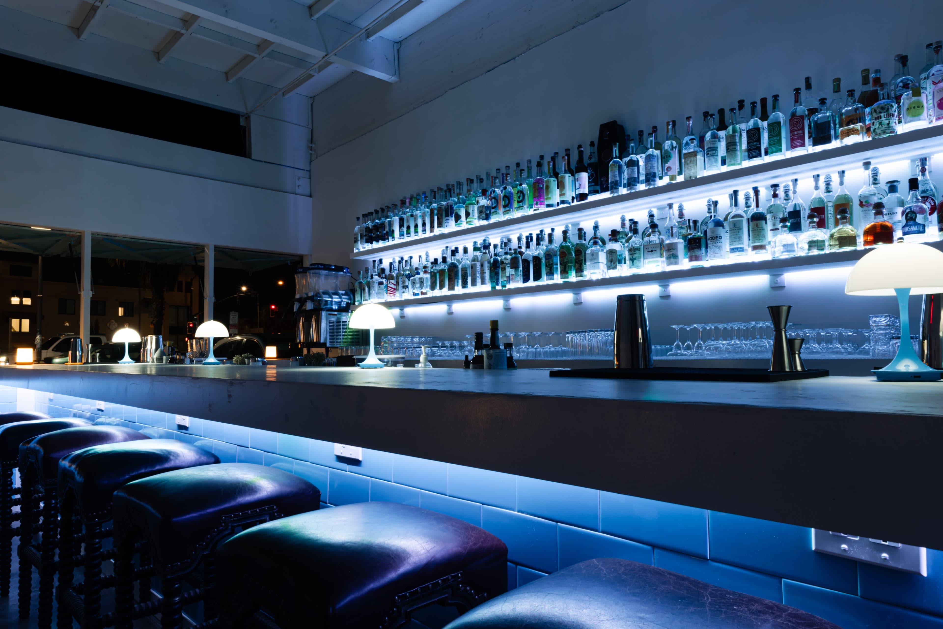 A bar with a long countertop displays an array of illuminated bottles on shelves above, accompanied by stylish, modern lighting.