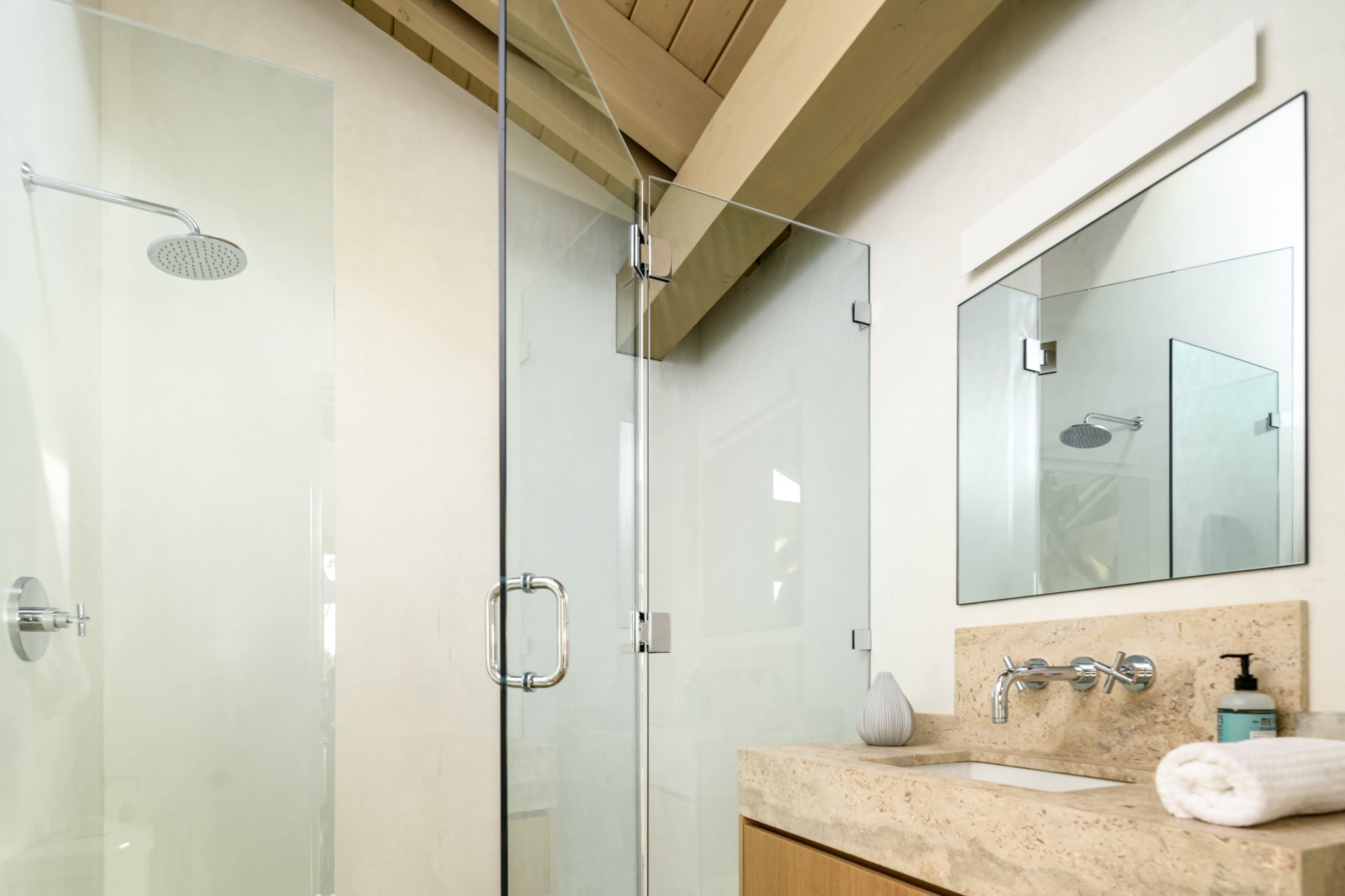 A modern bathroom features a glass shower enclosure, a stone countertop with a sink, and a large mirror, all under a sloped ceiling with wooden beams.