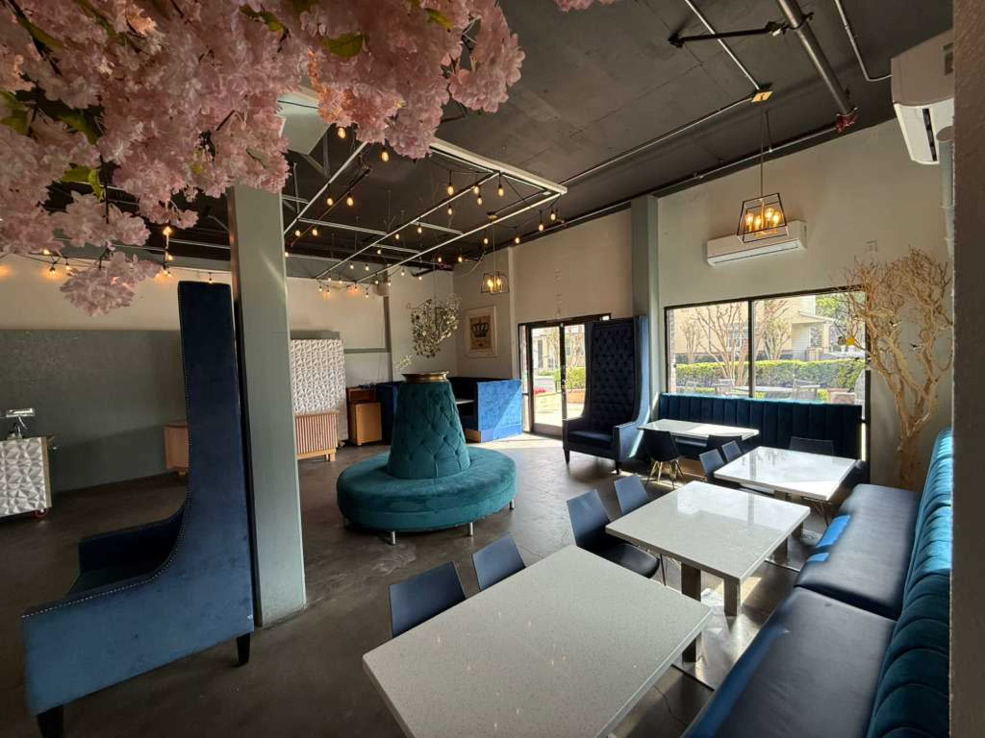 The image shows a modern interior of a restaurant or café featuring plush seating, a round seating area, and decorative cherry blossom branches.