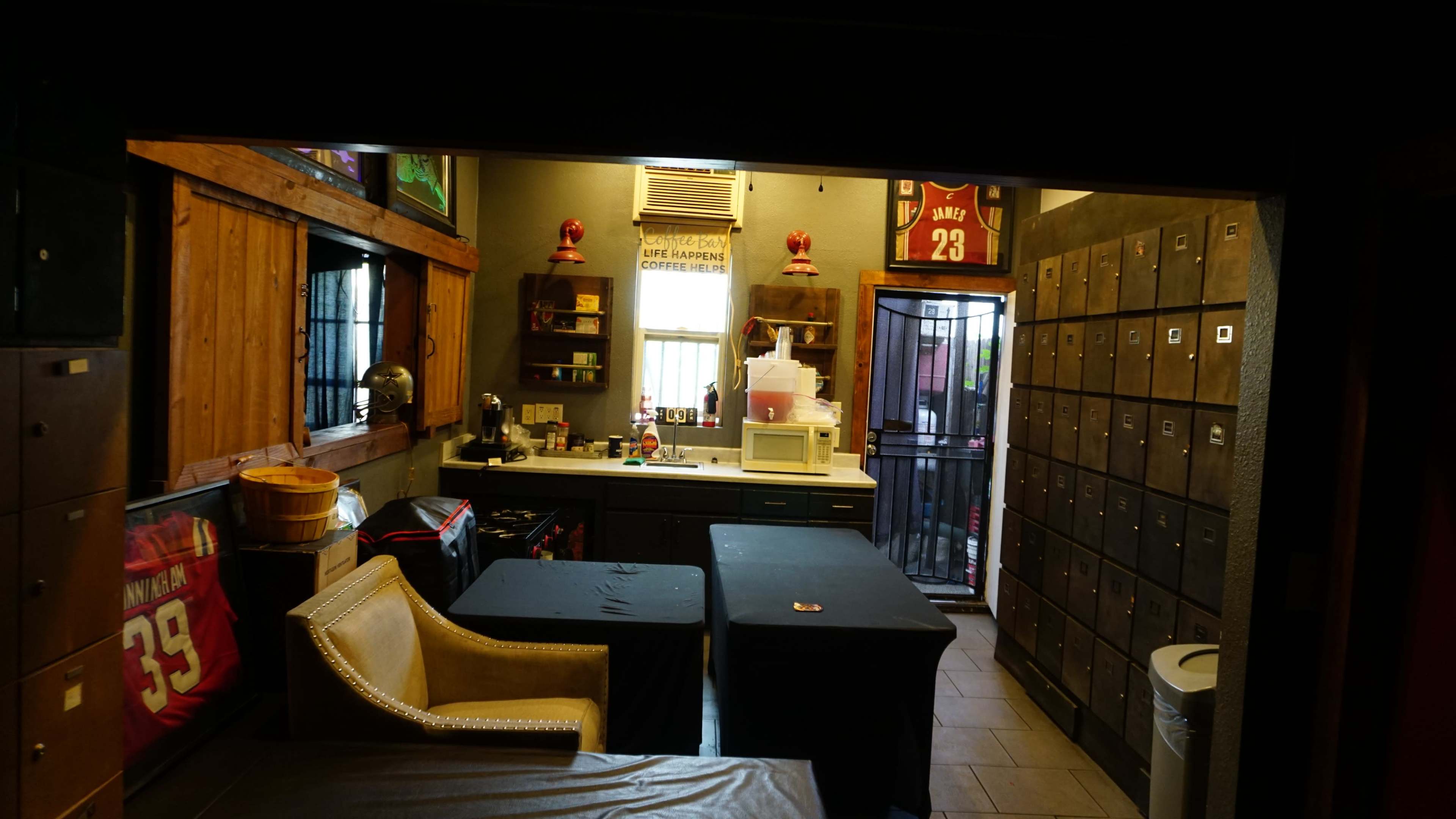 The image shows a small room with dark cabinets, a countertop, a microwave, and a few tables, decorated with sports jerseys and memorabilia.
