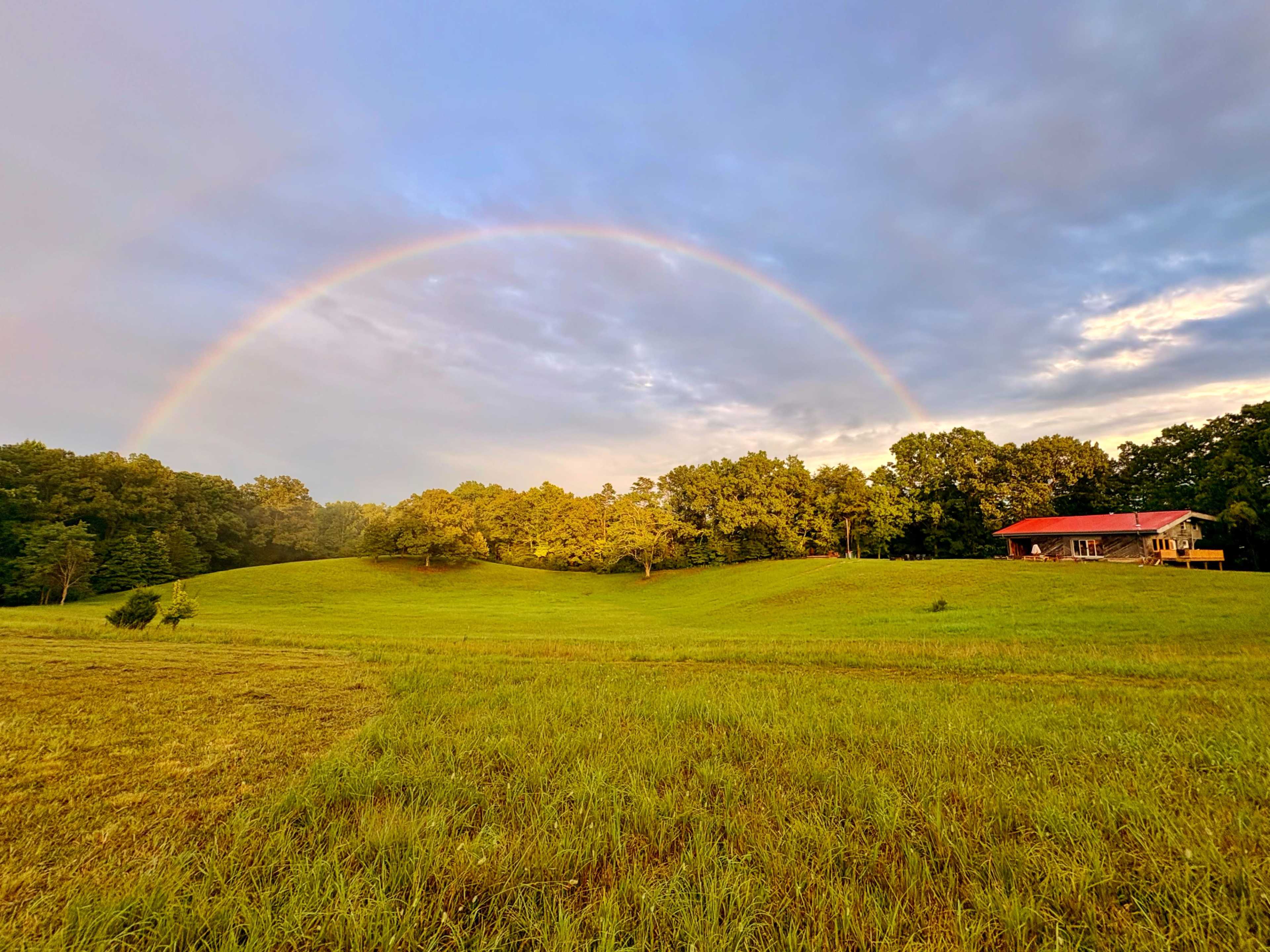 A rainbow arcs over a green hillside with a house nestled among trees in the distance.