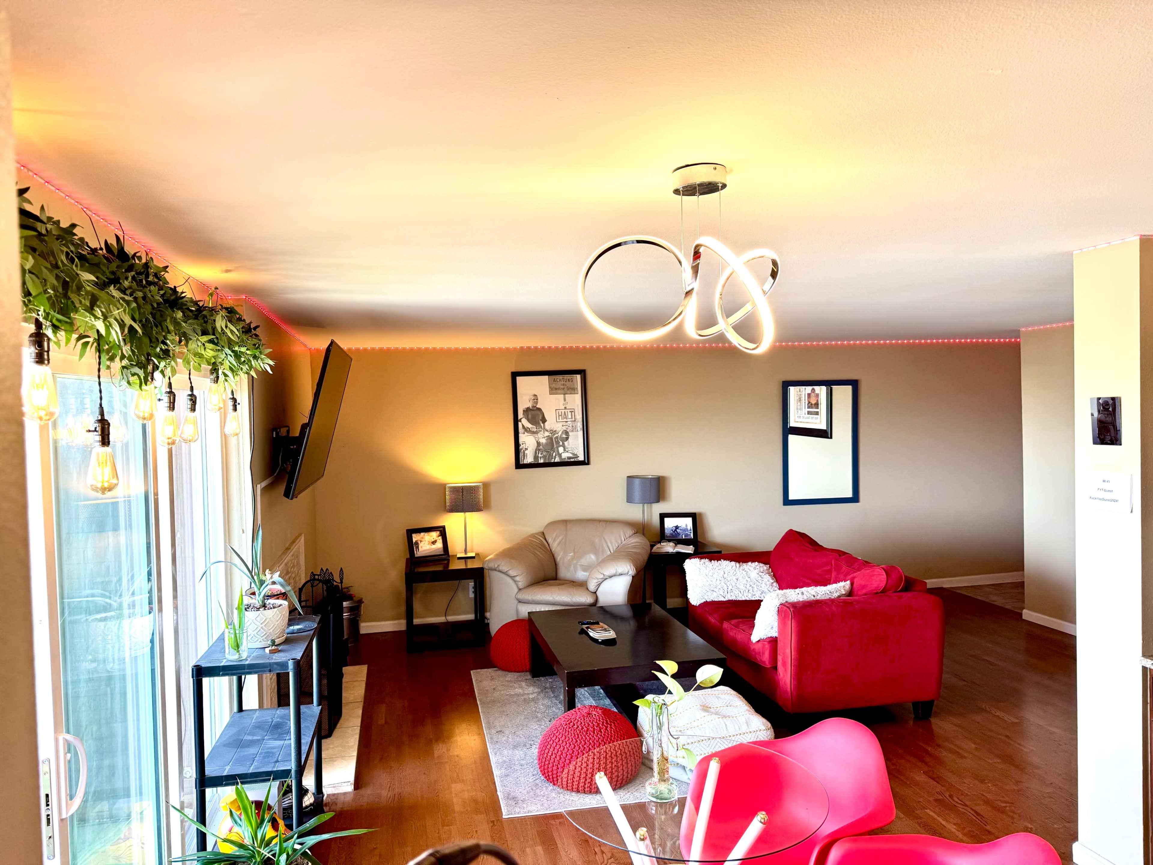 A modern living room features a red sectional sofa, a beige loveseat, a coffee table, and a television mounted on the wall, with ambient lighting along the ceiling.