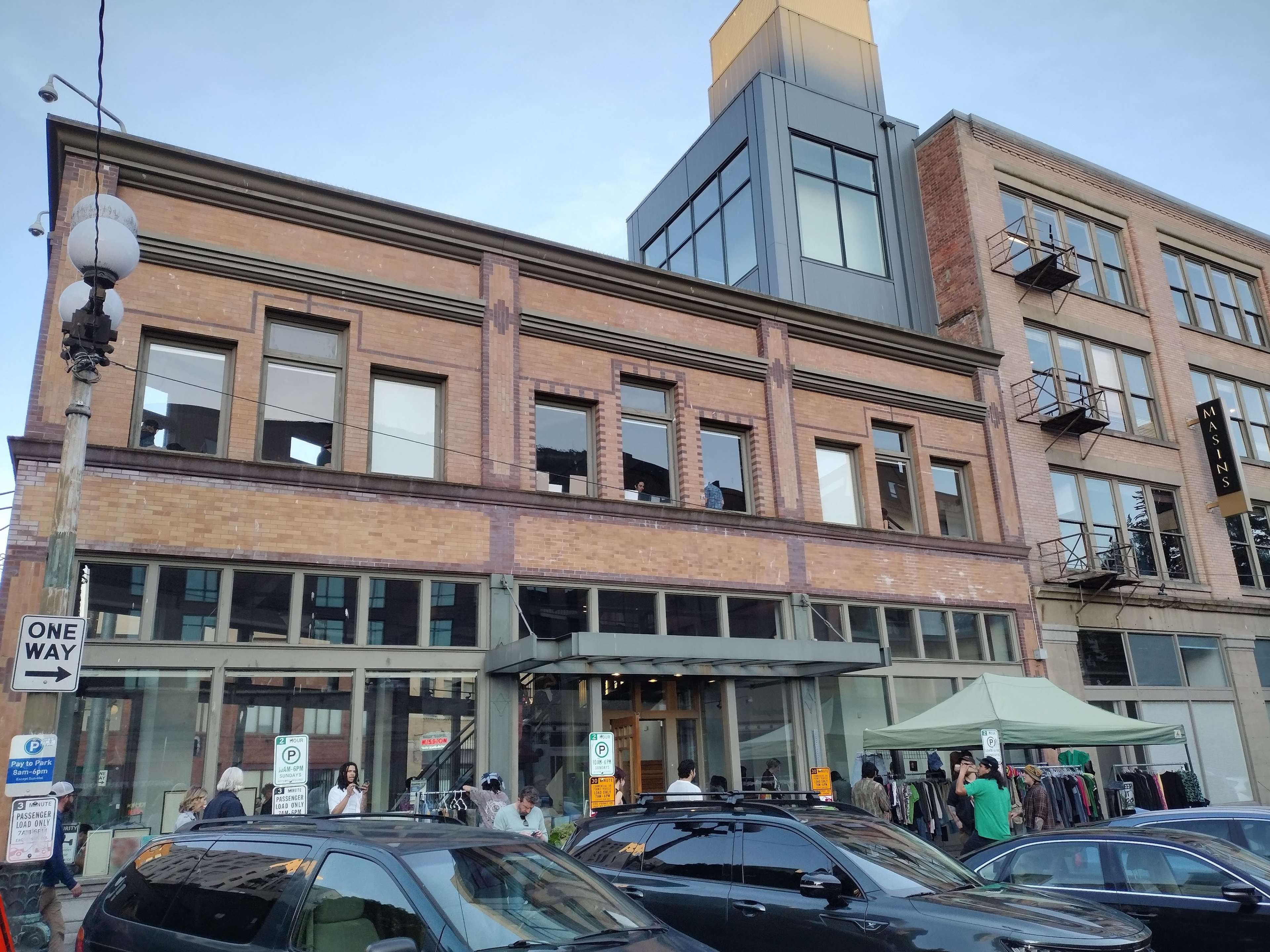 The image shows a three-story brick building with large windows and an outdoor clothing market in front of it, situated on a street with parked cars.