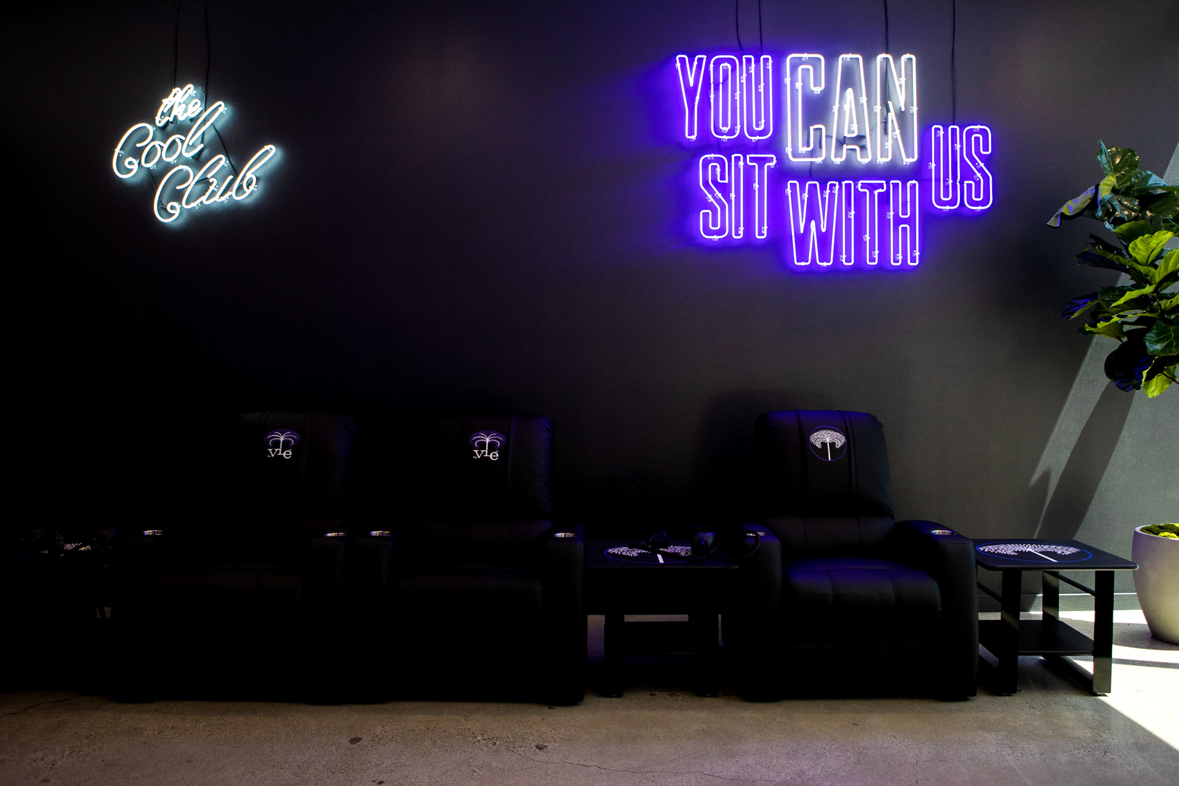 The image shows a modern lounge area featuring three black reclining chairs and neon signs that read "the Cool Club" and "YOU CAN SIT WITH US" against a dark wall.