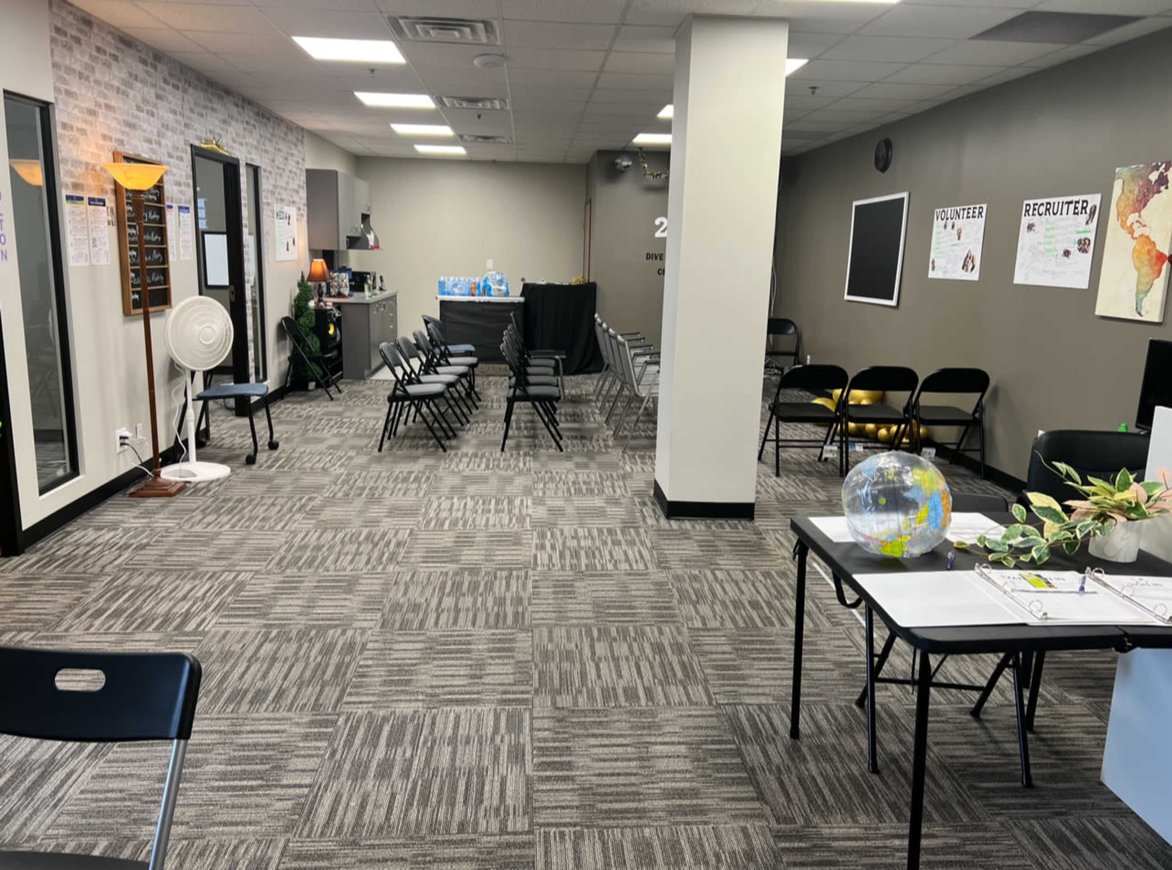 A spacious room with folding chairs arranged in rows, a reception desk at the back, and tables set up with various materials on the side.
