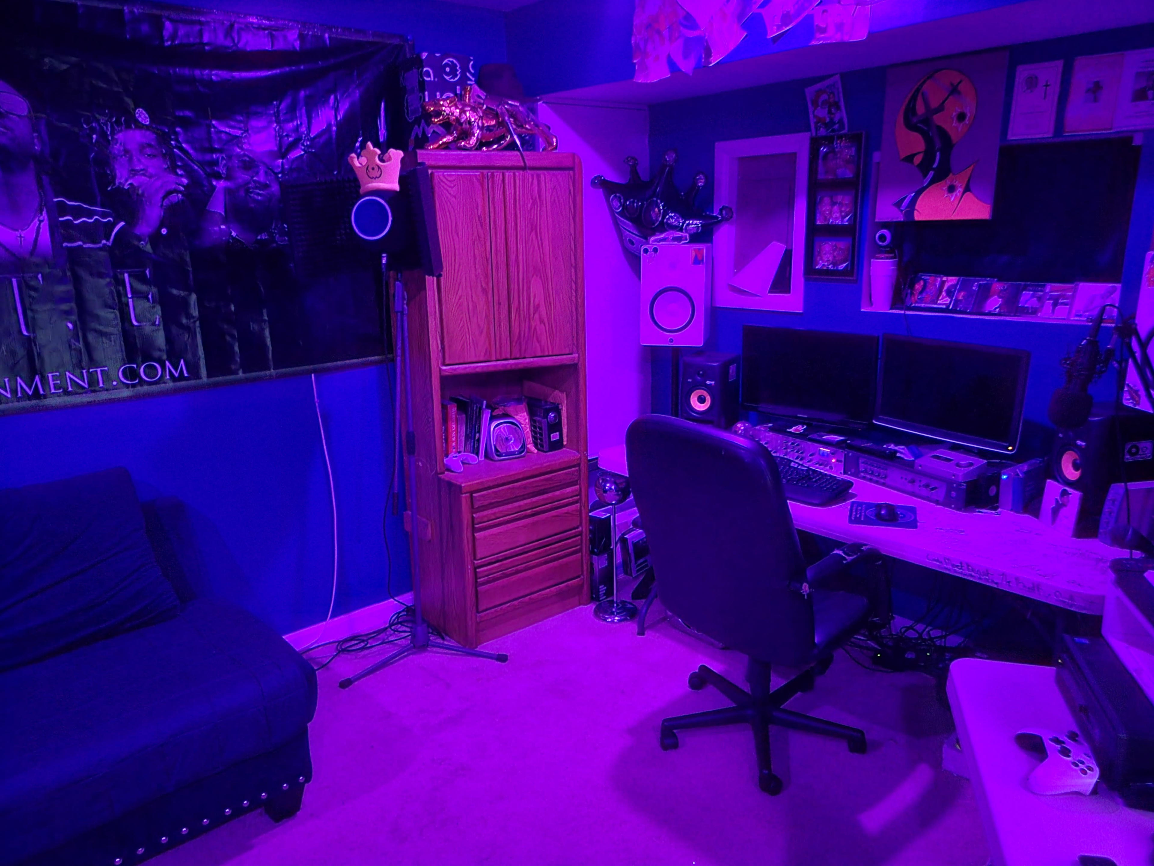A room set up as a recording studio, featuring a desk with dual monitors, audio equipment, a black swivel chair, and purple lighting.