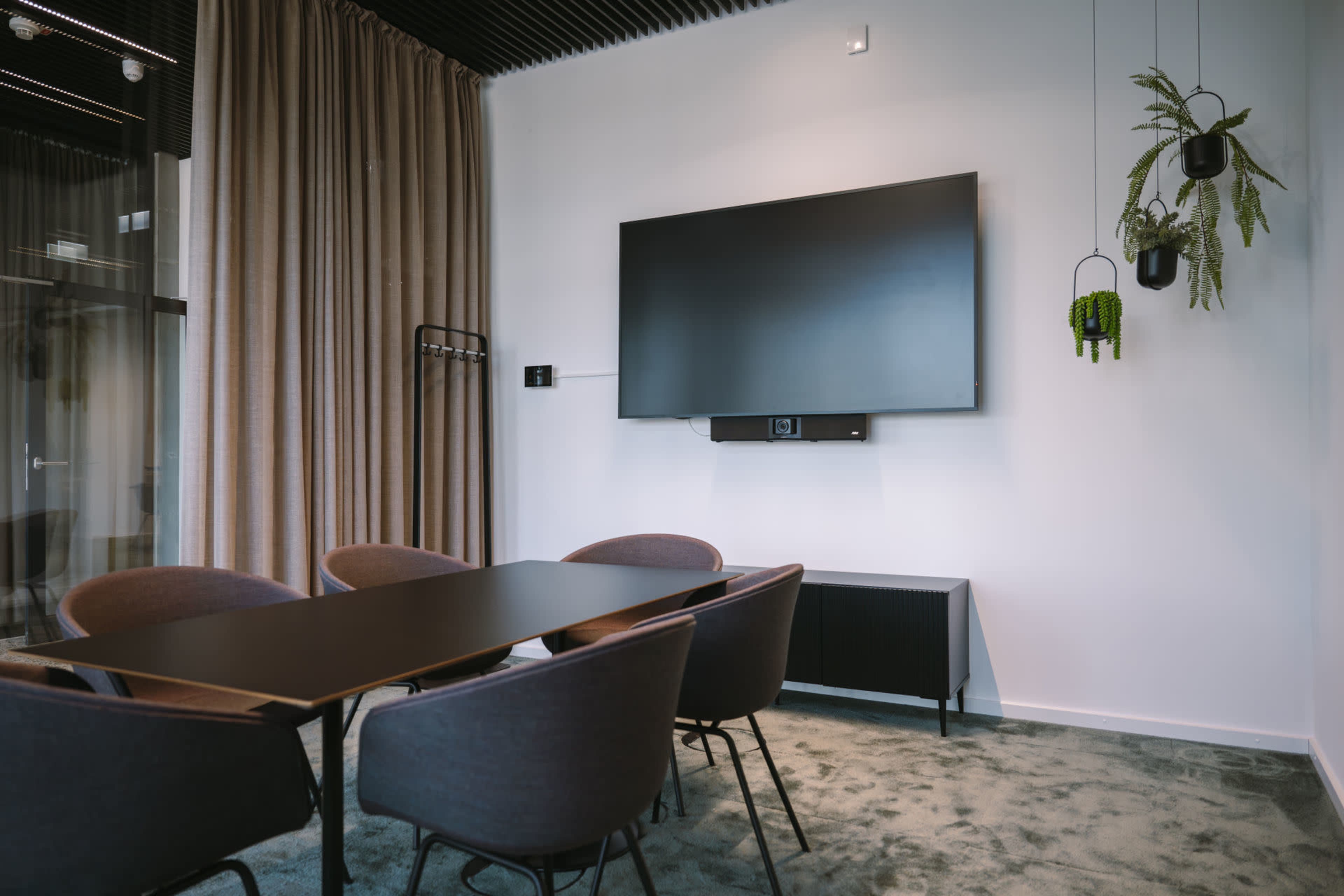 A modern conference room features a large wall-mounted TV, a rectangular black table, and upholstered chairs surrounding it, with curtains and wall-mounted plants in the background.
