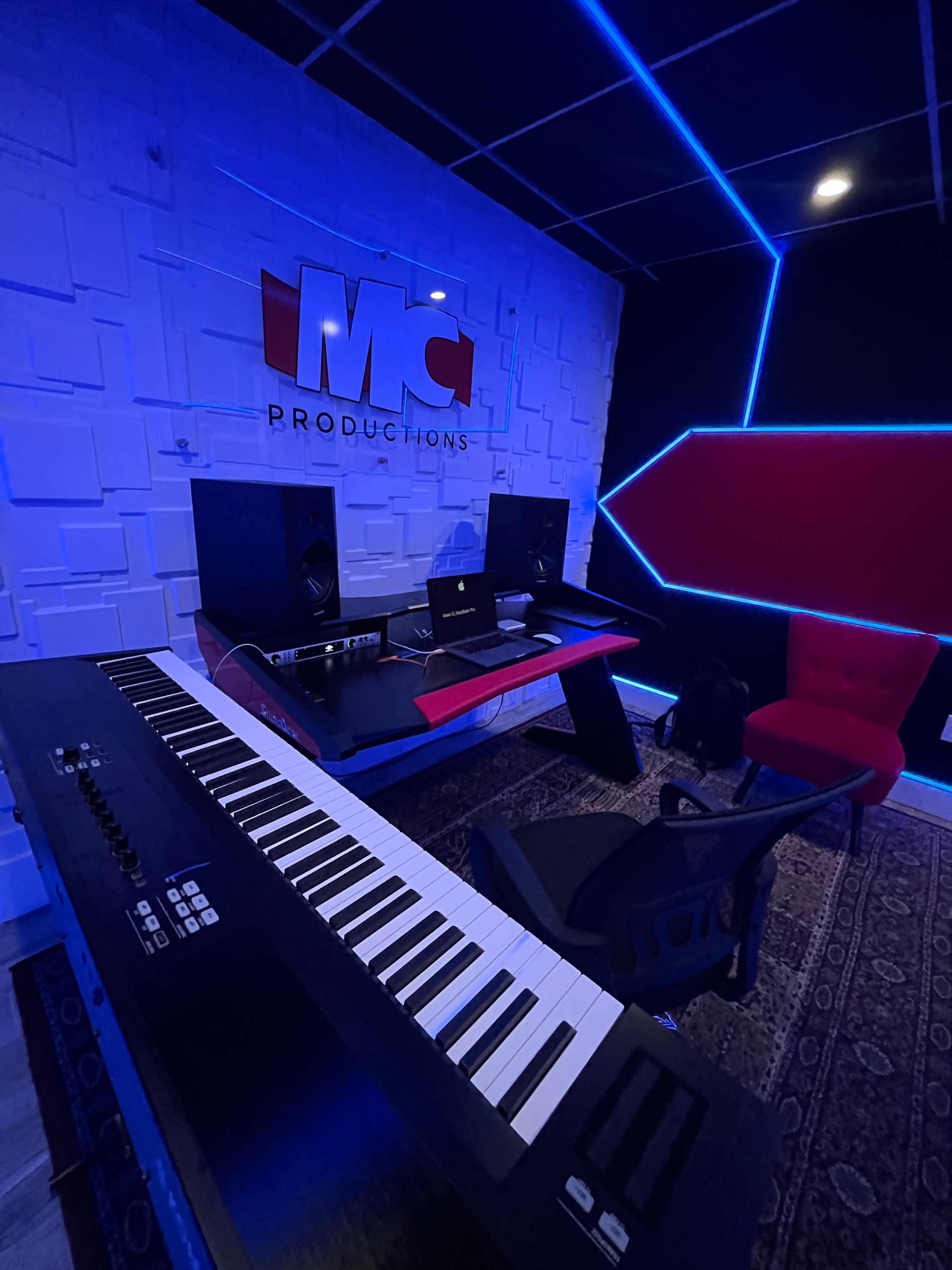 A modern music production studio features a keyboard, desk with a laptop, and sound equipment against a geometric backdrop with blue lighting.