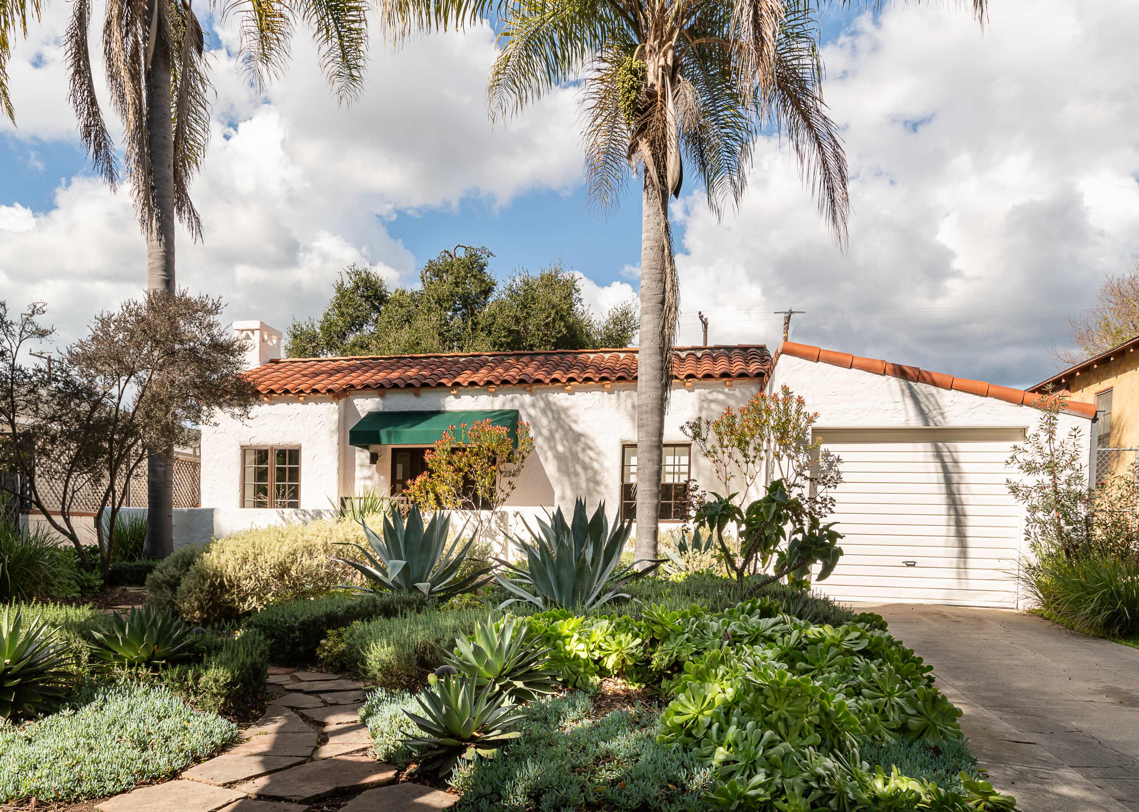 A single-story white house with a red tile roof is surrounded by various plants and a stone pathway, featuring a garage on the right.