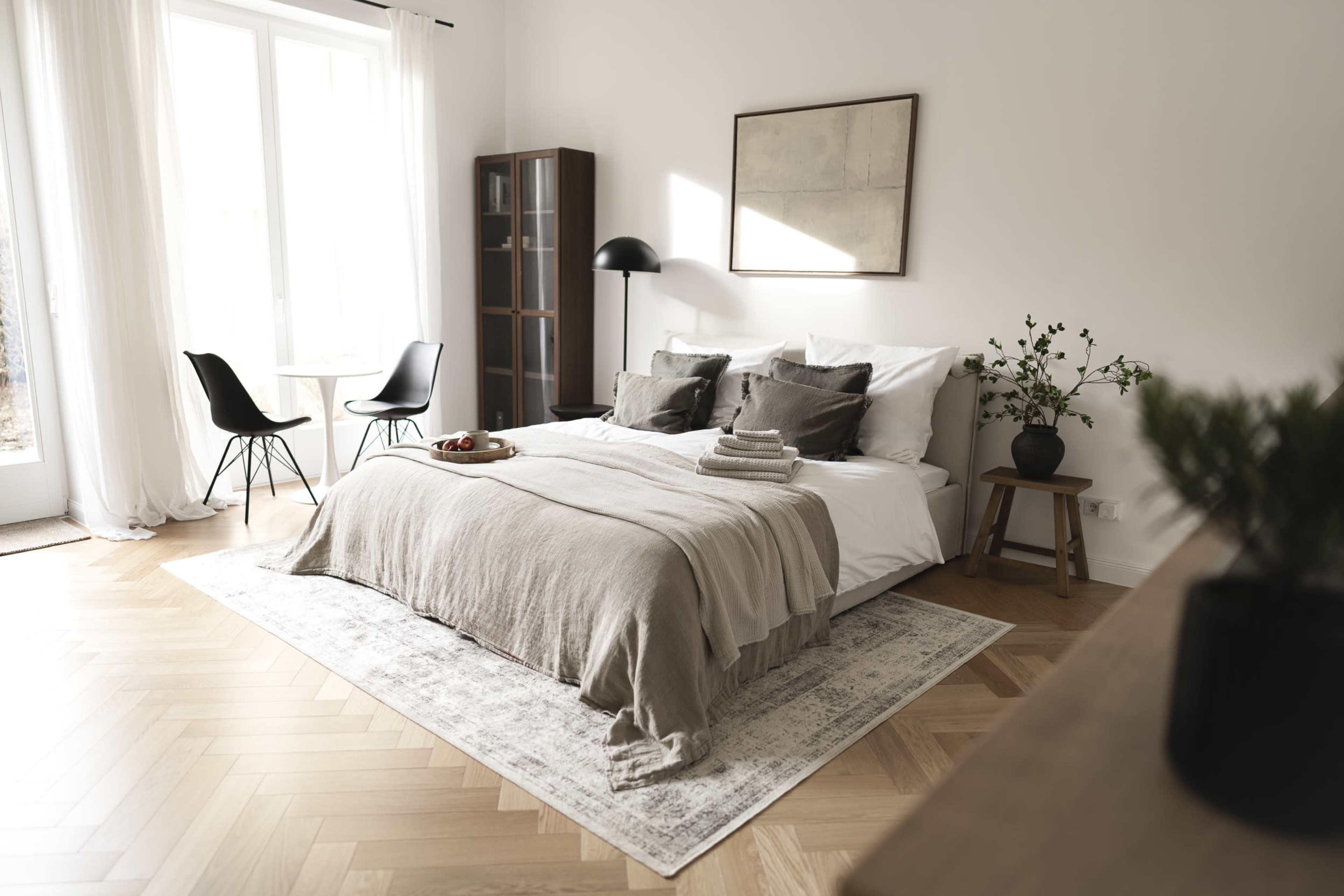 A neatly arranged bedroom features a large bed with soft linens, a small table with chairs, and natural light streaming through a window.