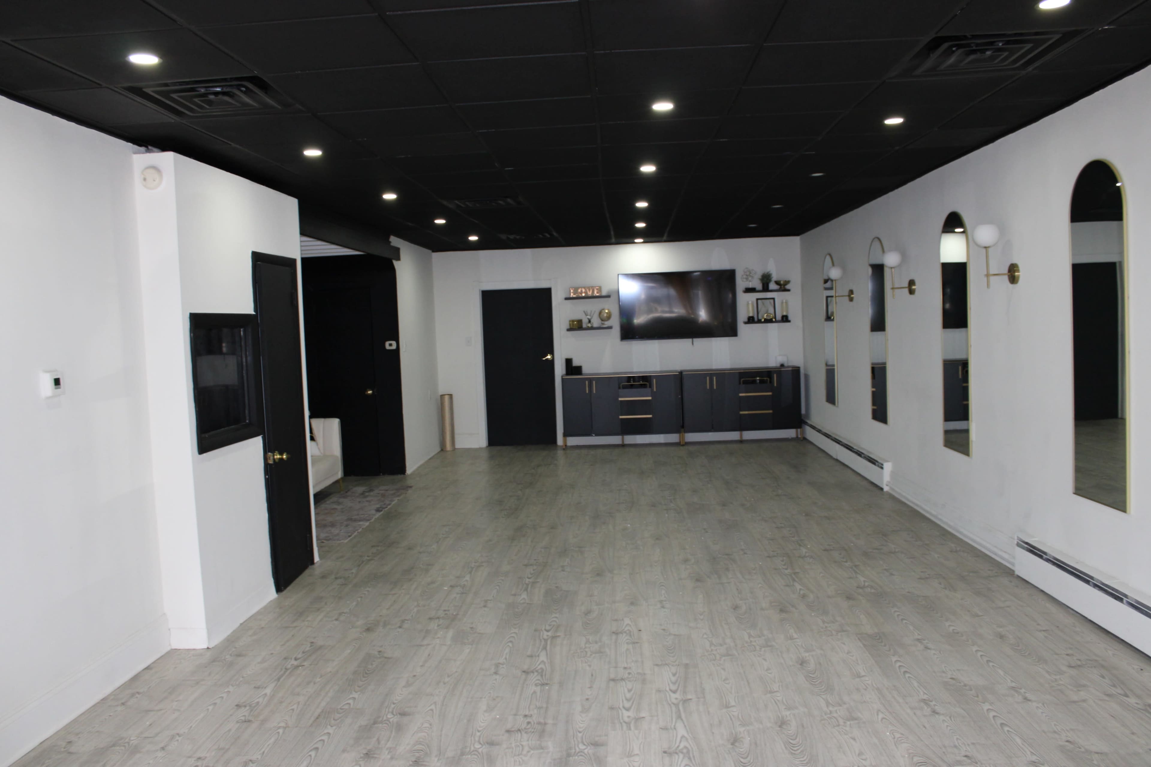 The image shows an empty room with a modern design featuring wooden flooring, a television mounted on the wall, and several decorative mirrors.