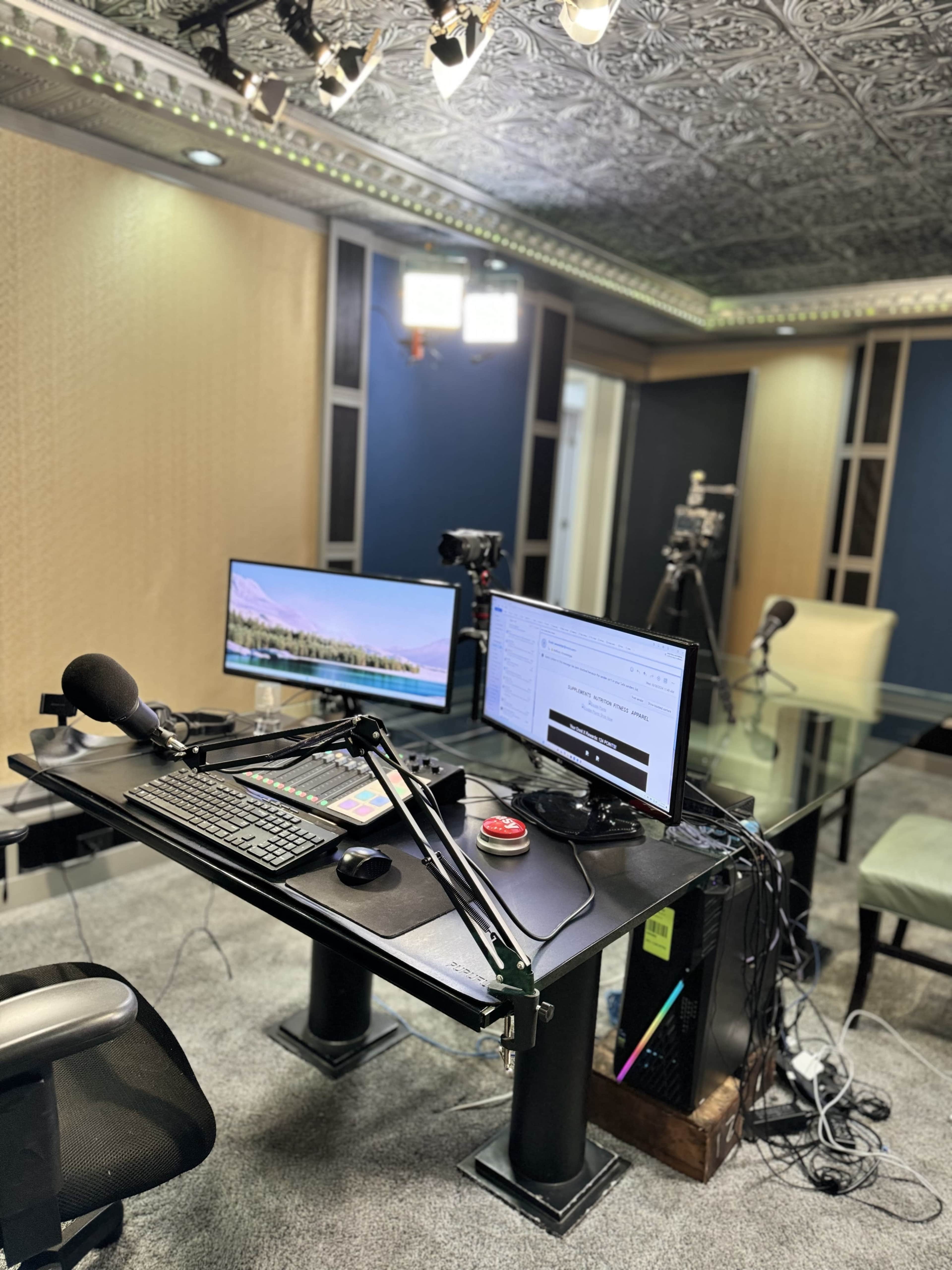 A modern podcasting or streaming setup is shown, featuring a desk with a keyboard, microphone on an arm, dual monitors, and various camera equipment in a well-lit room.