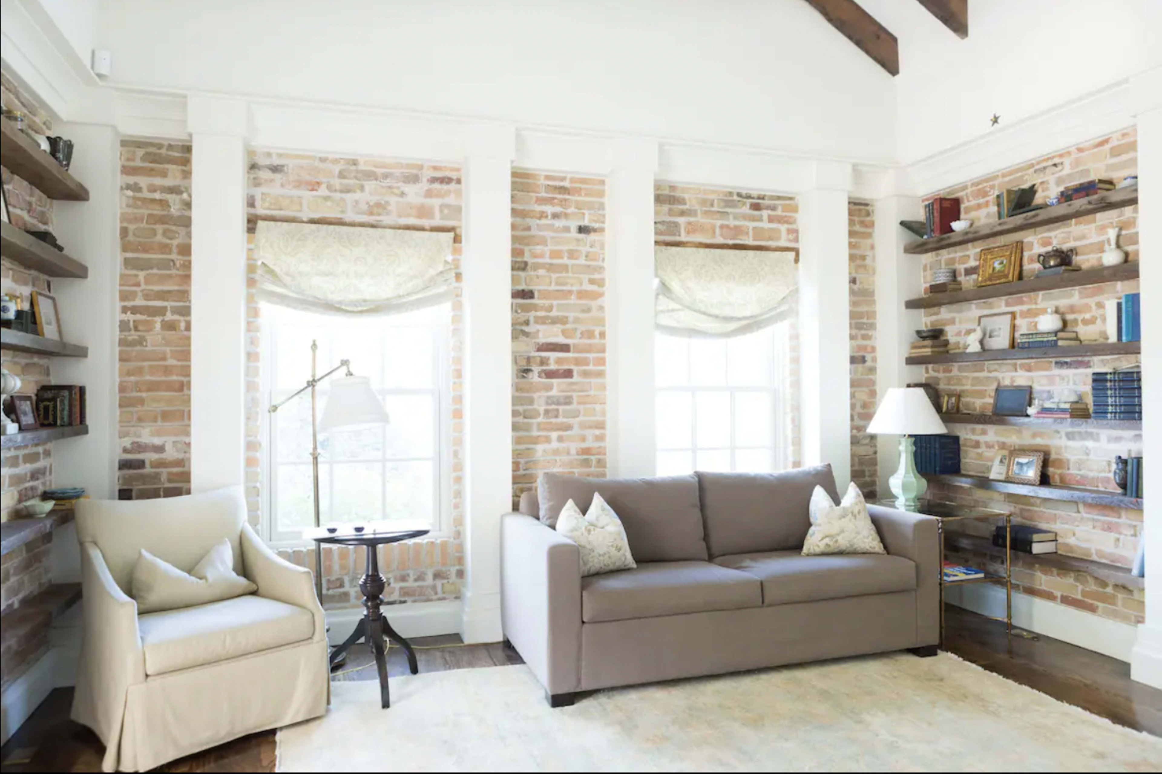 A living room with exposed brick walls, a light-colored sofa, an armchair, a side table, and bookshelves filled with various items.