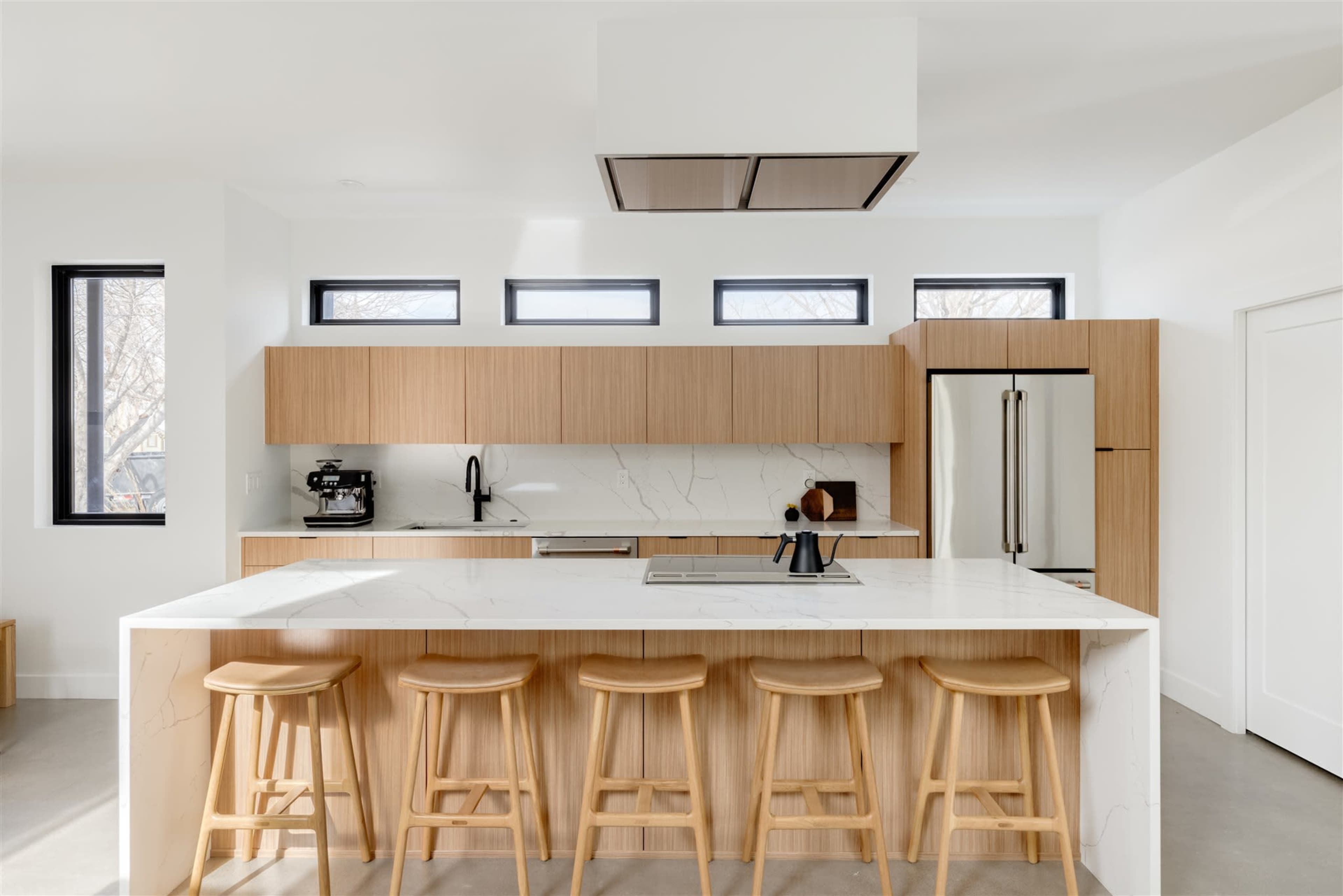 A modern kitchen features wooden cabinetry, a marble countertop island with seating, and large windows allowing natural light.