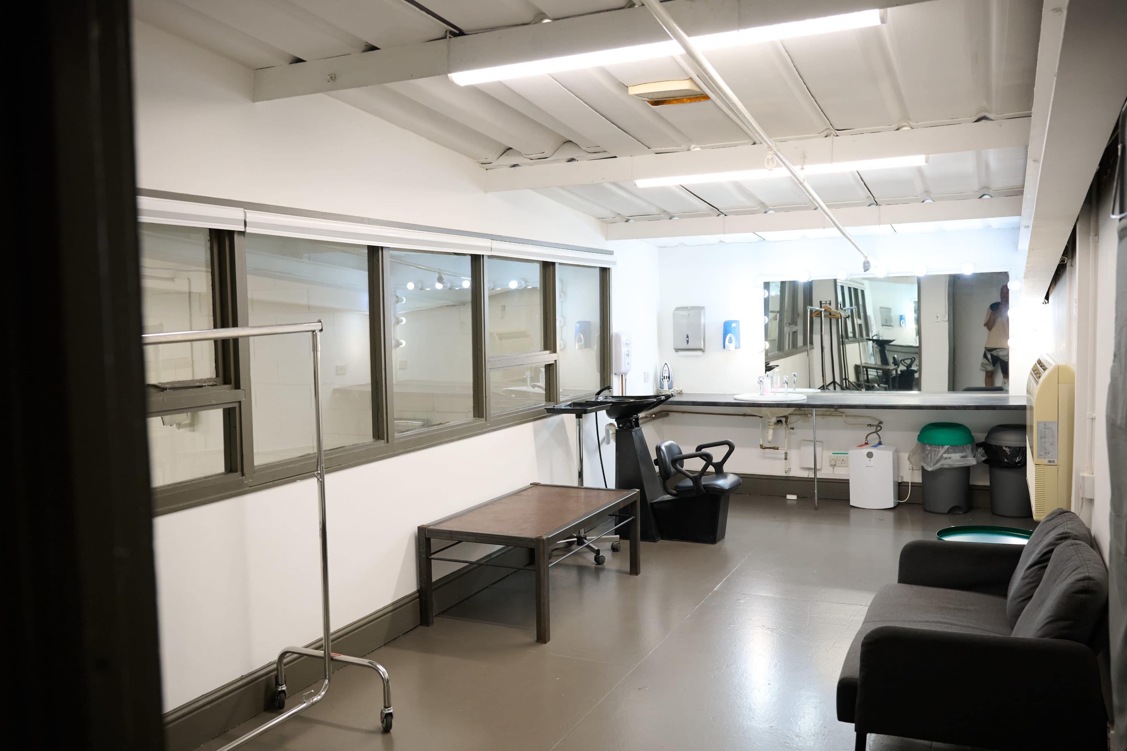 The image shows a minimalistic room with a table, a chair, a clothing rack, and a mirror along one wall, featuring large windows that provide natural light.