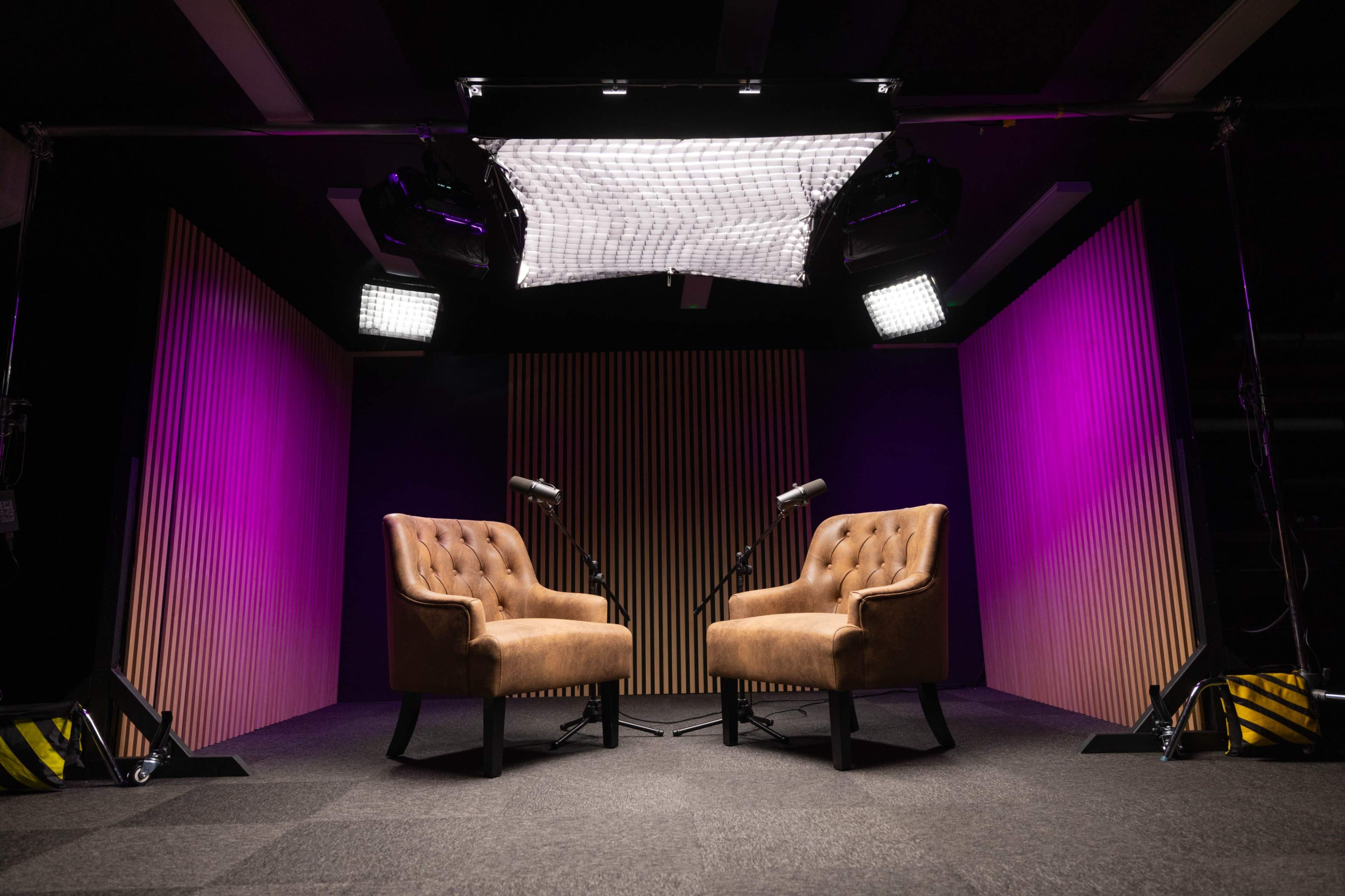 The image shows two brown tufted chairs positioned facing each other under bright studio lights, set against a backdrop with vertical wooden slats and purple lighting.