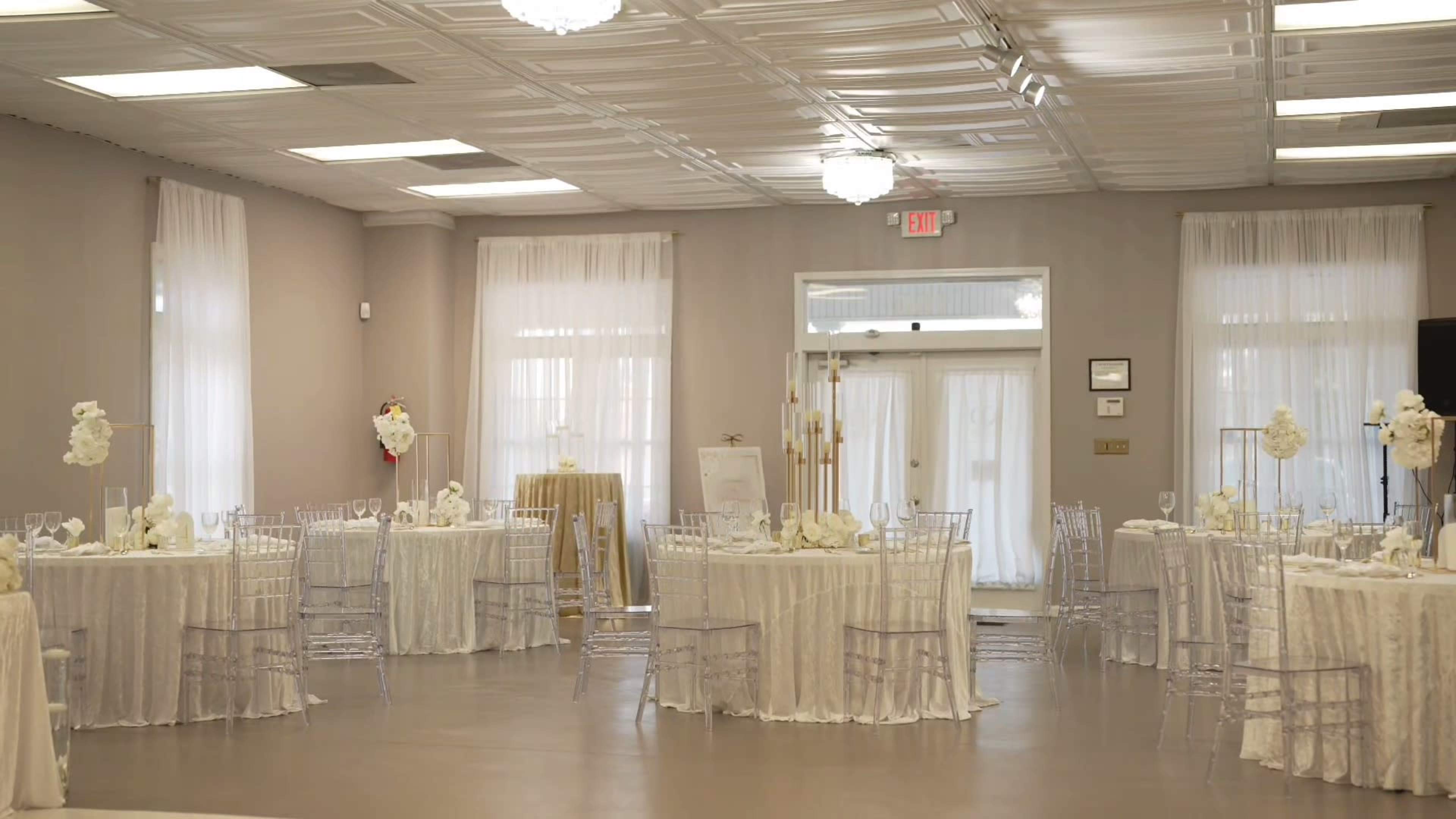 An elegantly arranged event space features round tables draped in white tablecloths, surrounded by clear acrylic chairs, with soft lighting from chandeliers above.