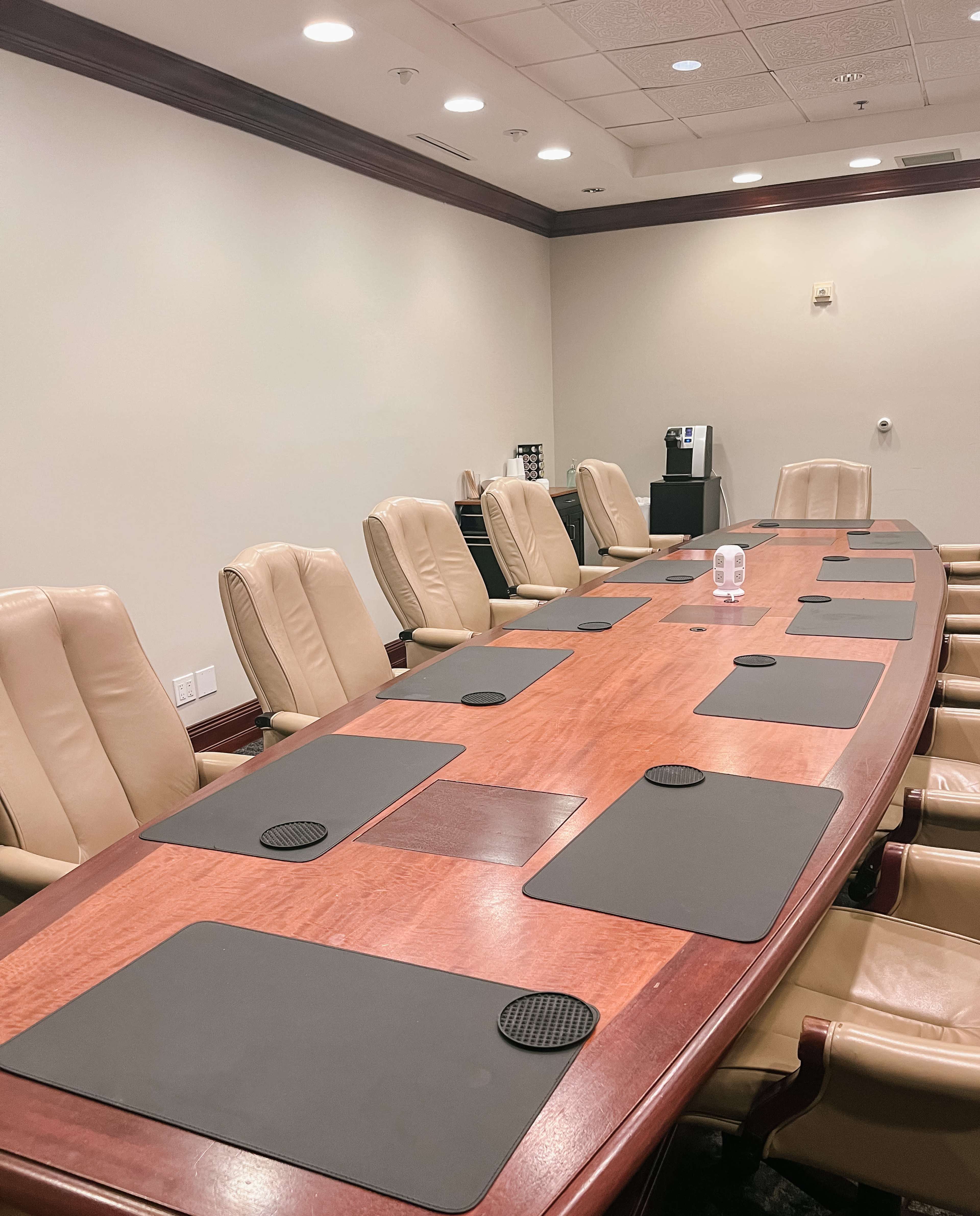 A large conference room features a wooden table surrounded by beige leather chairs, with black placemats placed before each seat.
