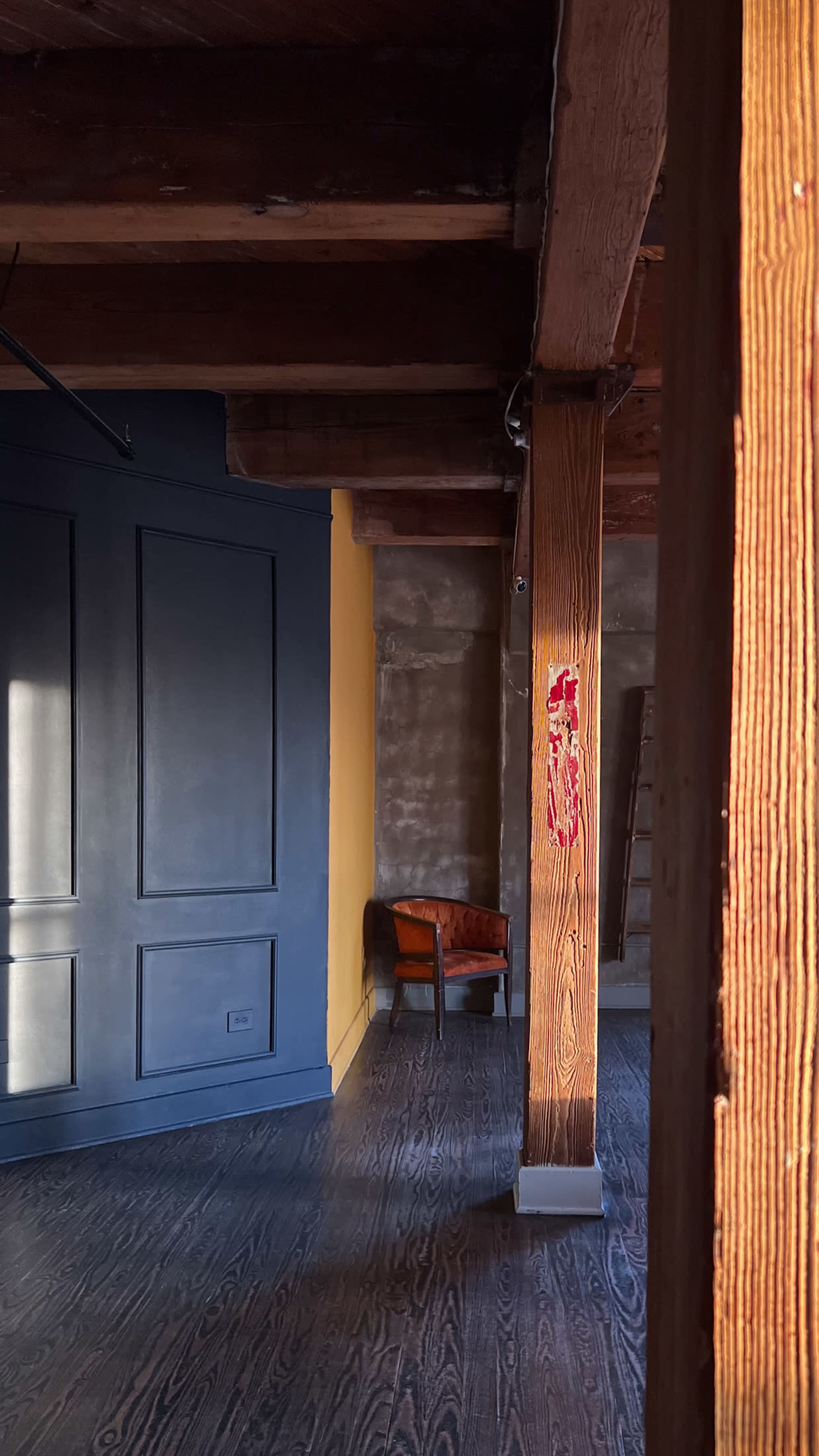 A partially furnished room with wooden beams, a yellow wall, and a brown chair in the corner.