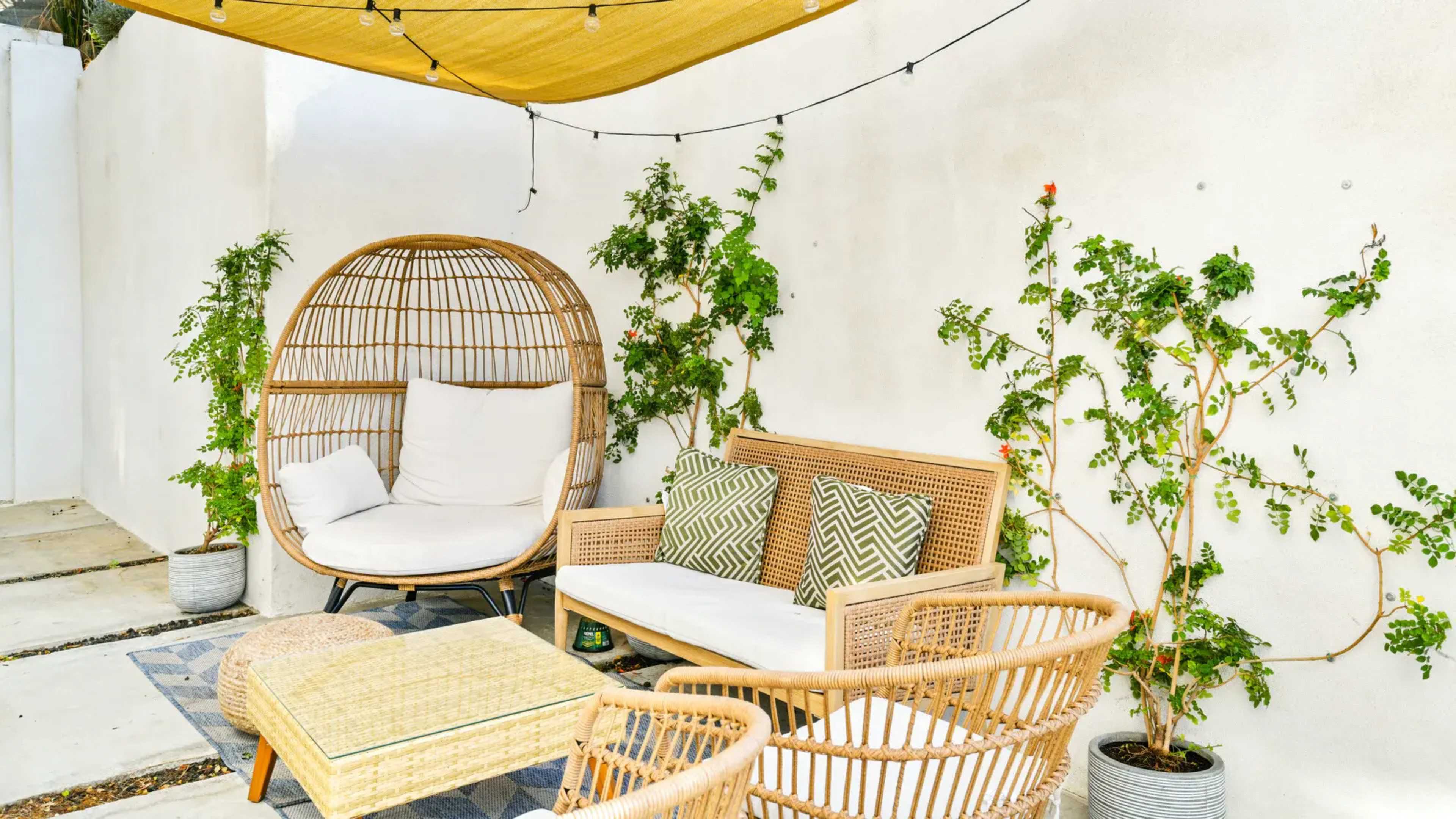 The scene features a cozy outdoor seating area with a circular rattan chair, a cushioned sofa, and potted plants against a white wall, all under a yellow canopy and string lights.