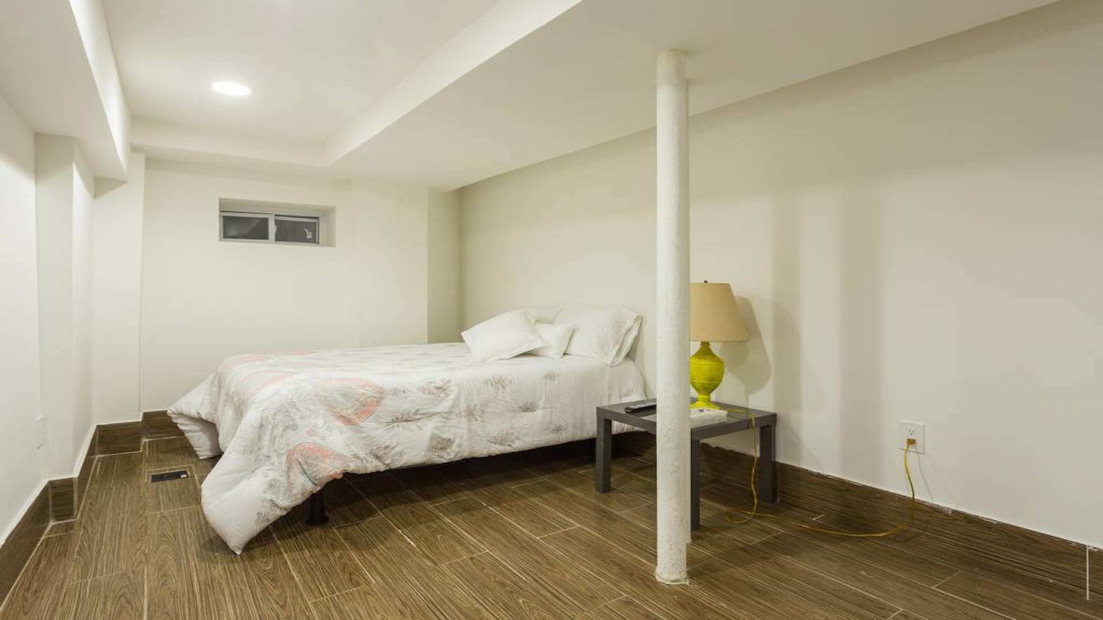 A small, minimalist bedroom features a bed with white bedding, a side table with a yellow lamp, and a central support column.