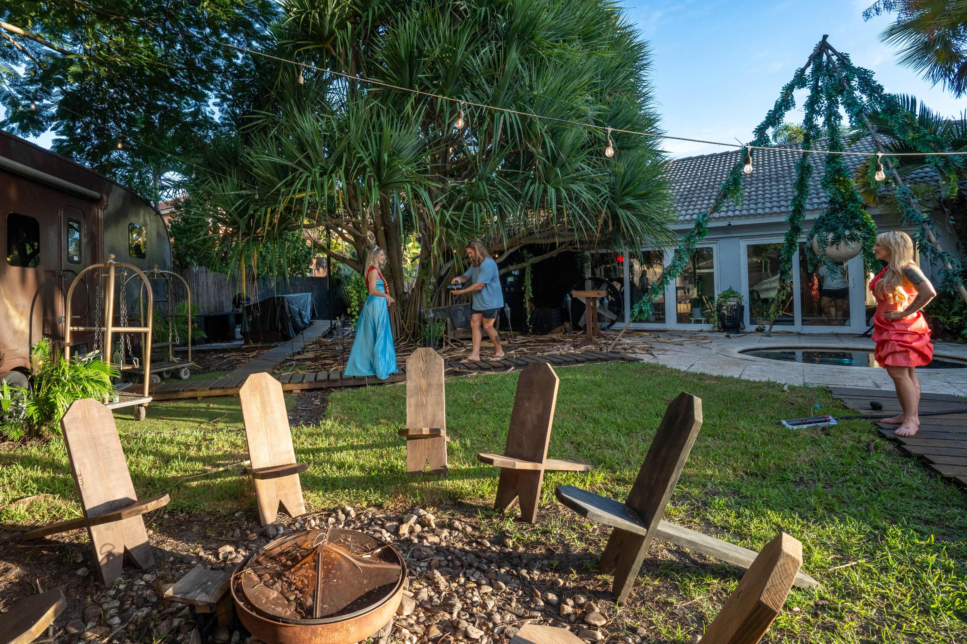 A backyard is set up with wooden chairs arranged around a fire pit, while a group of people gathers near a large tree, with a house visible in the background.