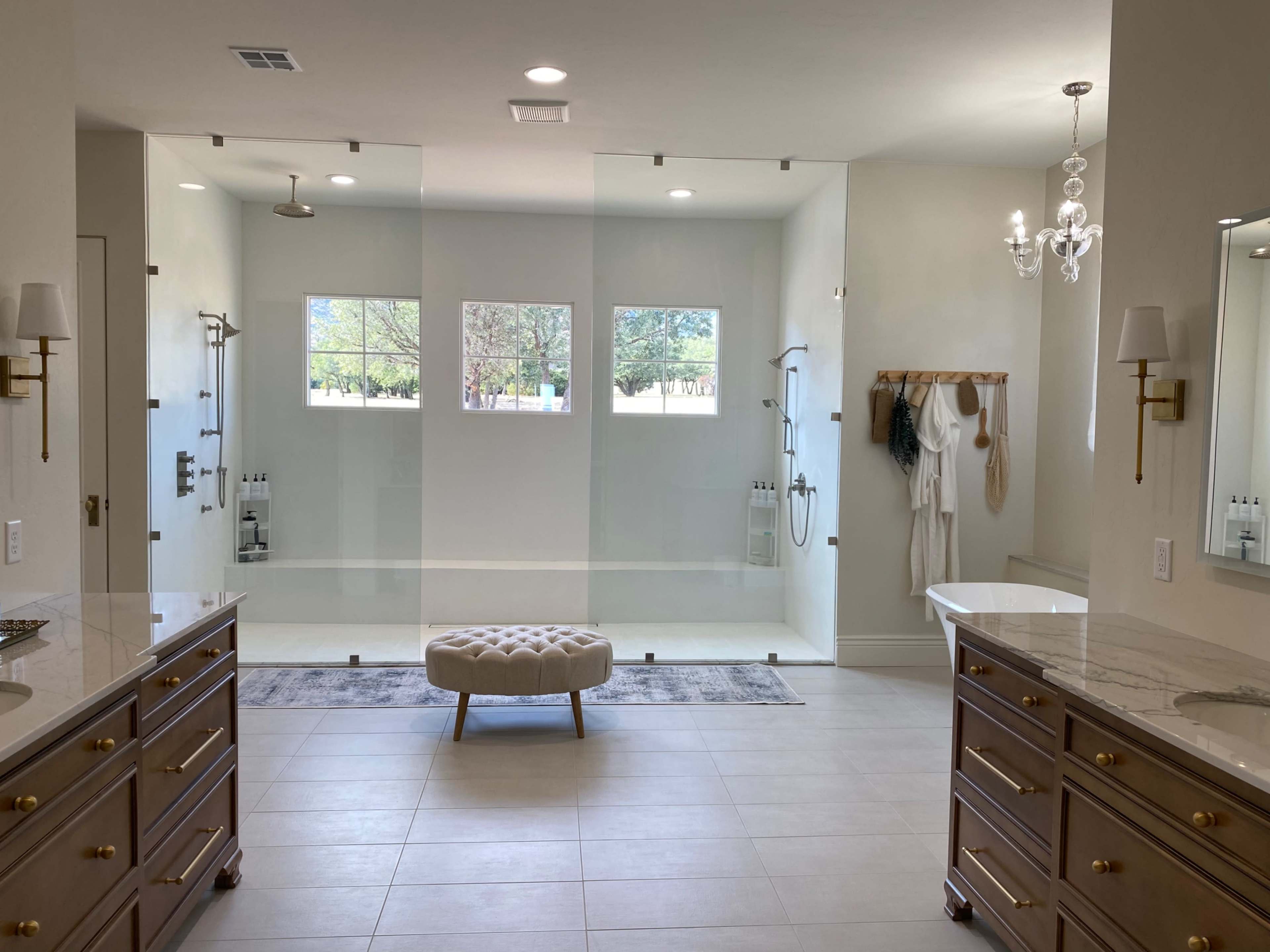 A modern bathroom featuring dual vanities, a central upholstered bench, and a large glass-enclosed shower area with multiple showerheads and viewable windows.