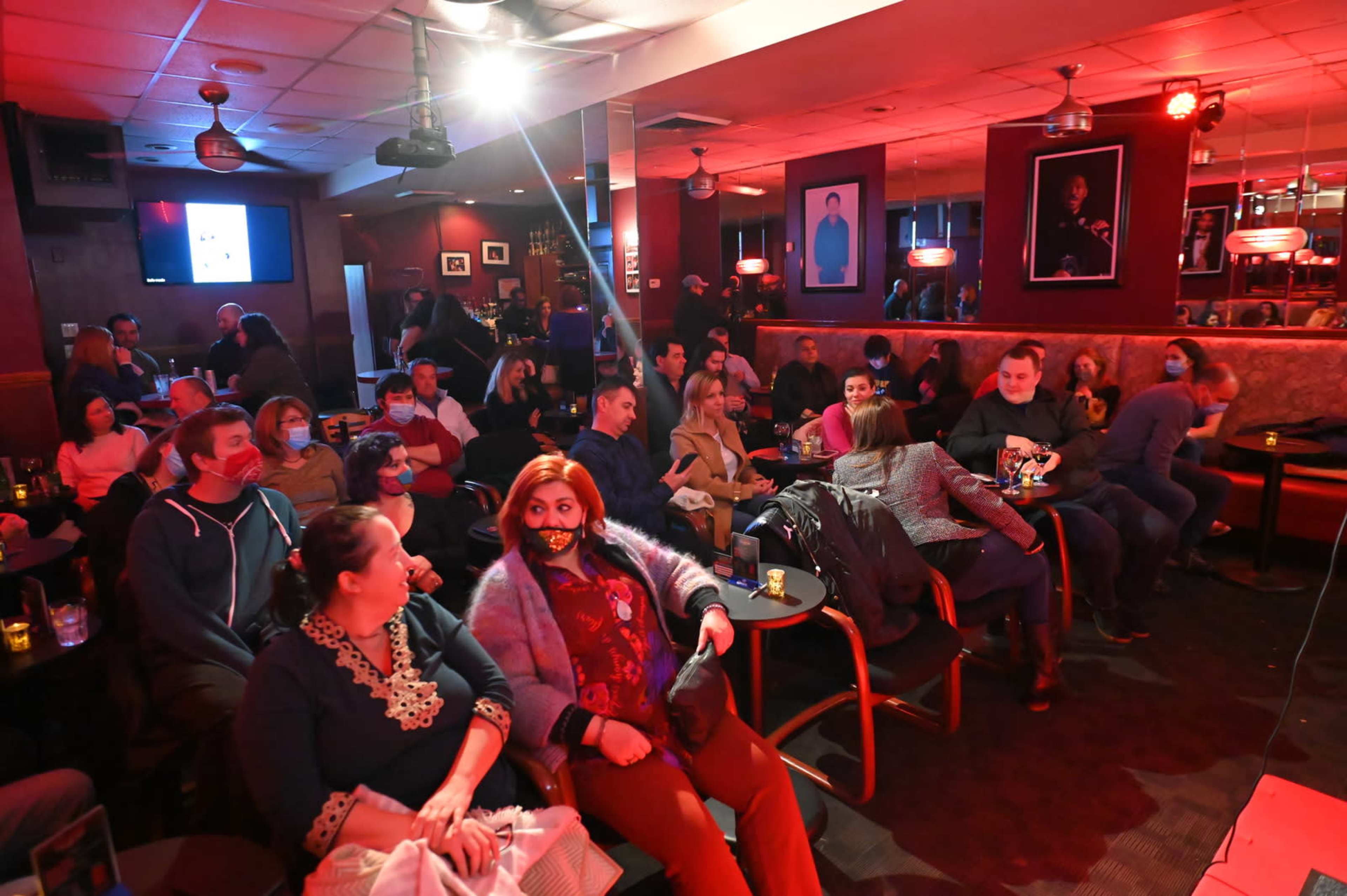 A crowd of people wearing masks sits in a dimly lit bar, with red lighting and a stage visible in the background.