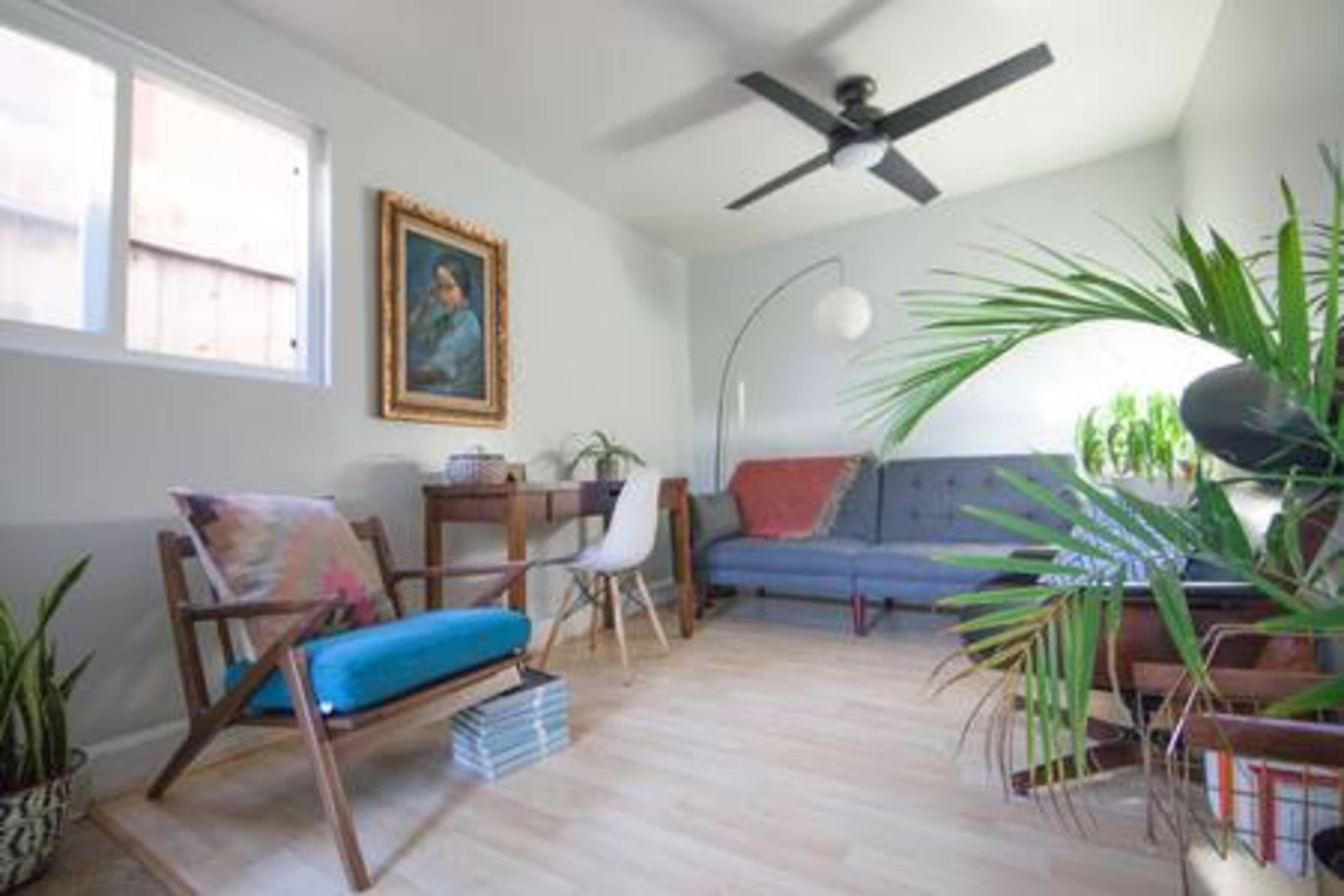 The image shows a well-lit living room featuring a gray sofa, a wooden chair with a blue cushion, a floor lamp, and several potted plants.