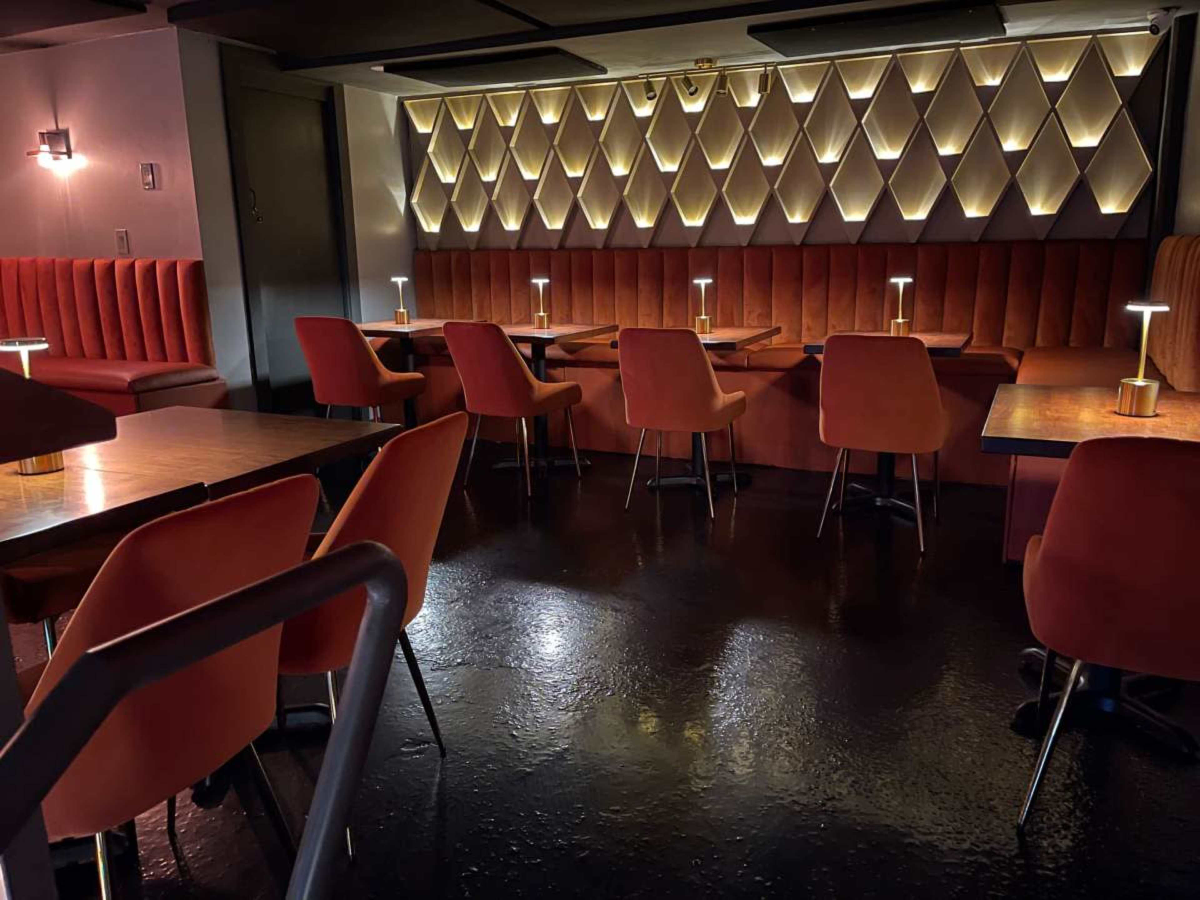 The image shows a dimly lit interior of a restaurant featuring orange upholstered chairs and wooden tables arranged in a cozy layout.