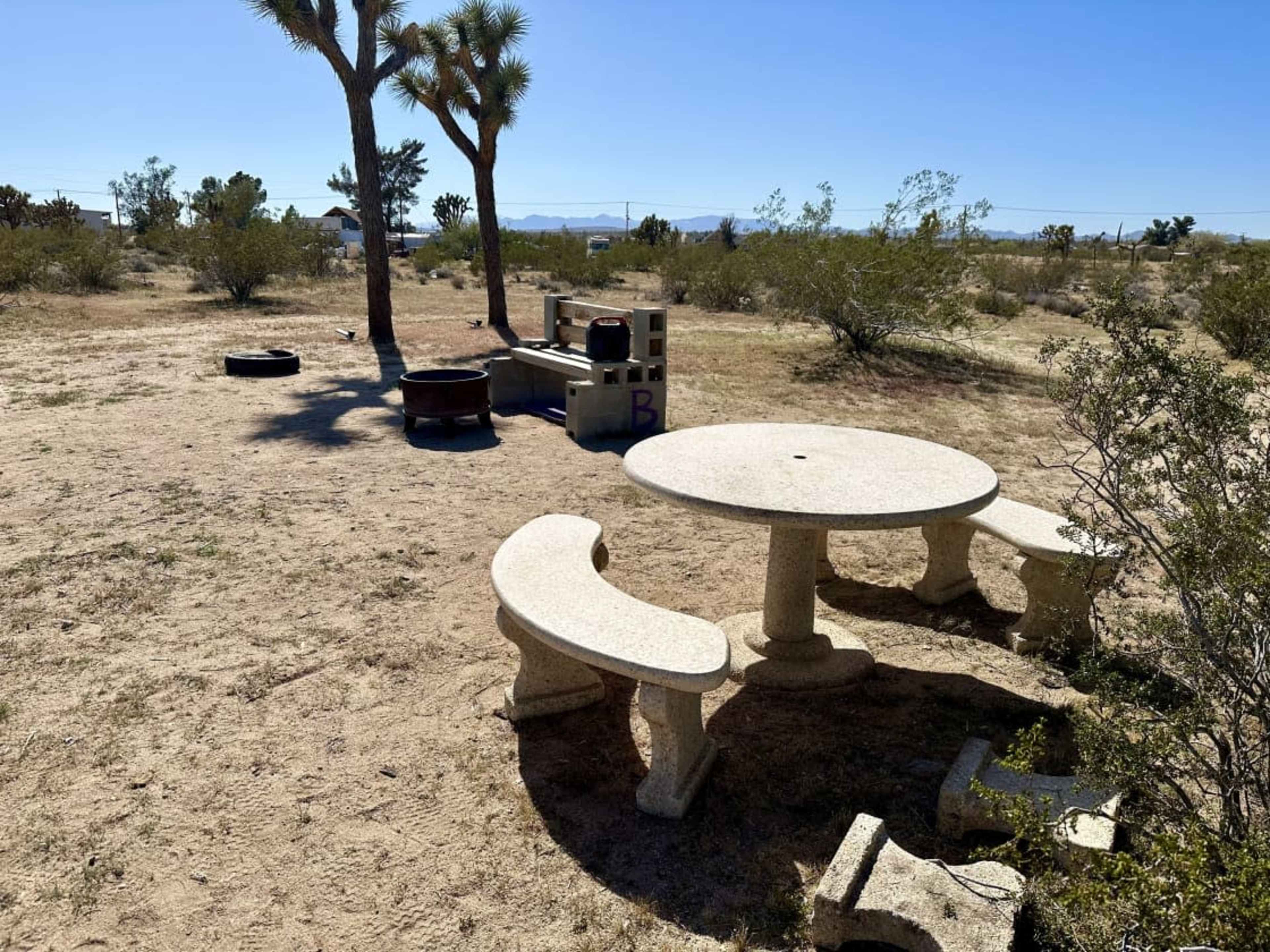 A circular stone table with two matching benches is situated in a desert landscape featuring sparse vegetation and distant mountains.