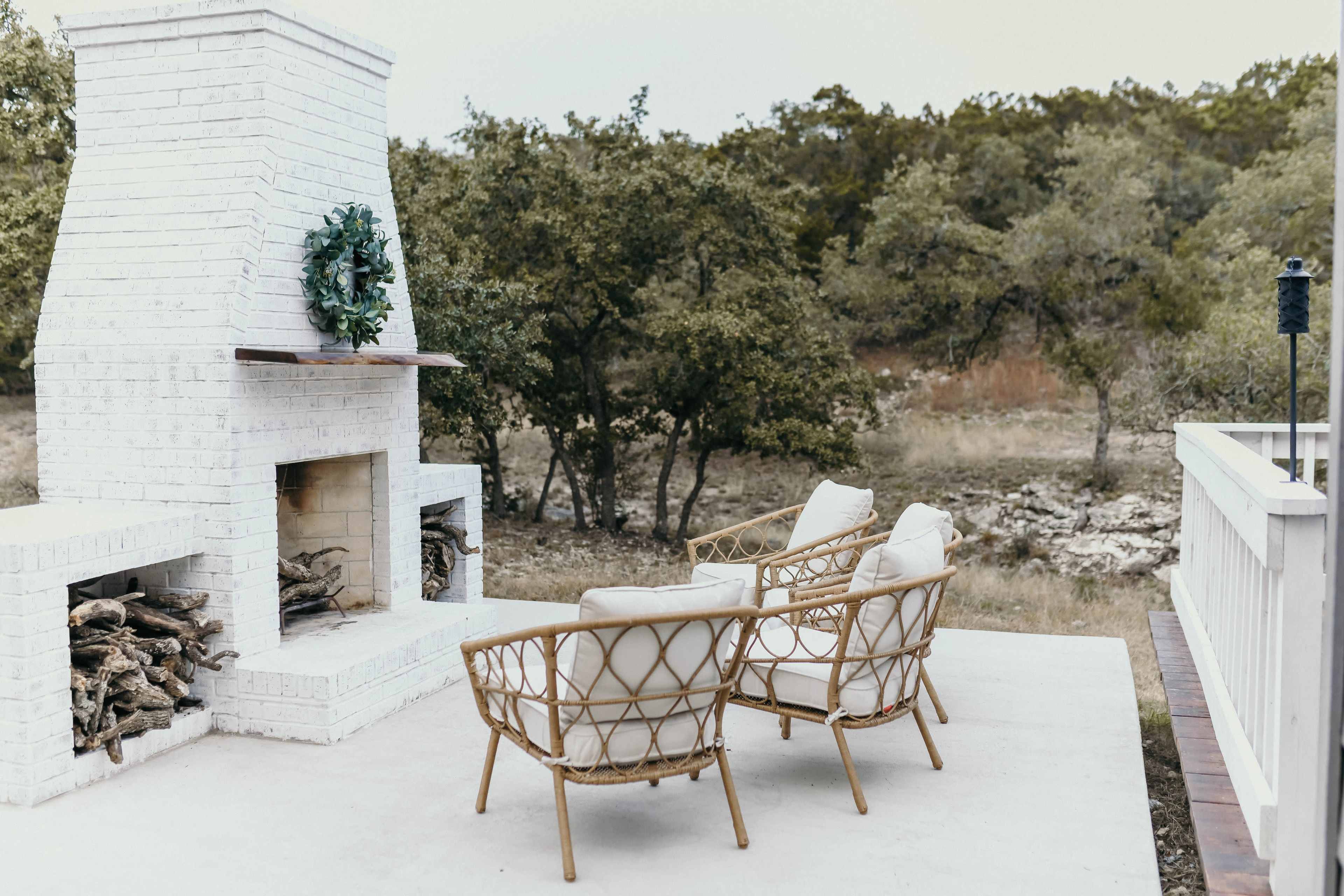 A white outdoor fireplace with a wreath sits between two rattan chairs on a patio overlooking a wooded area.