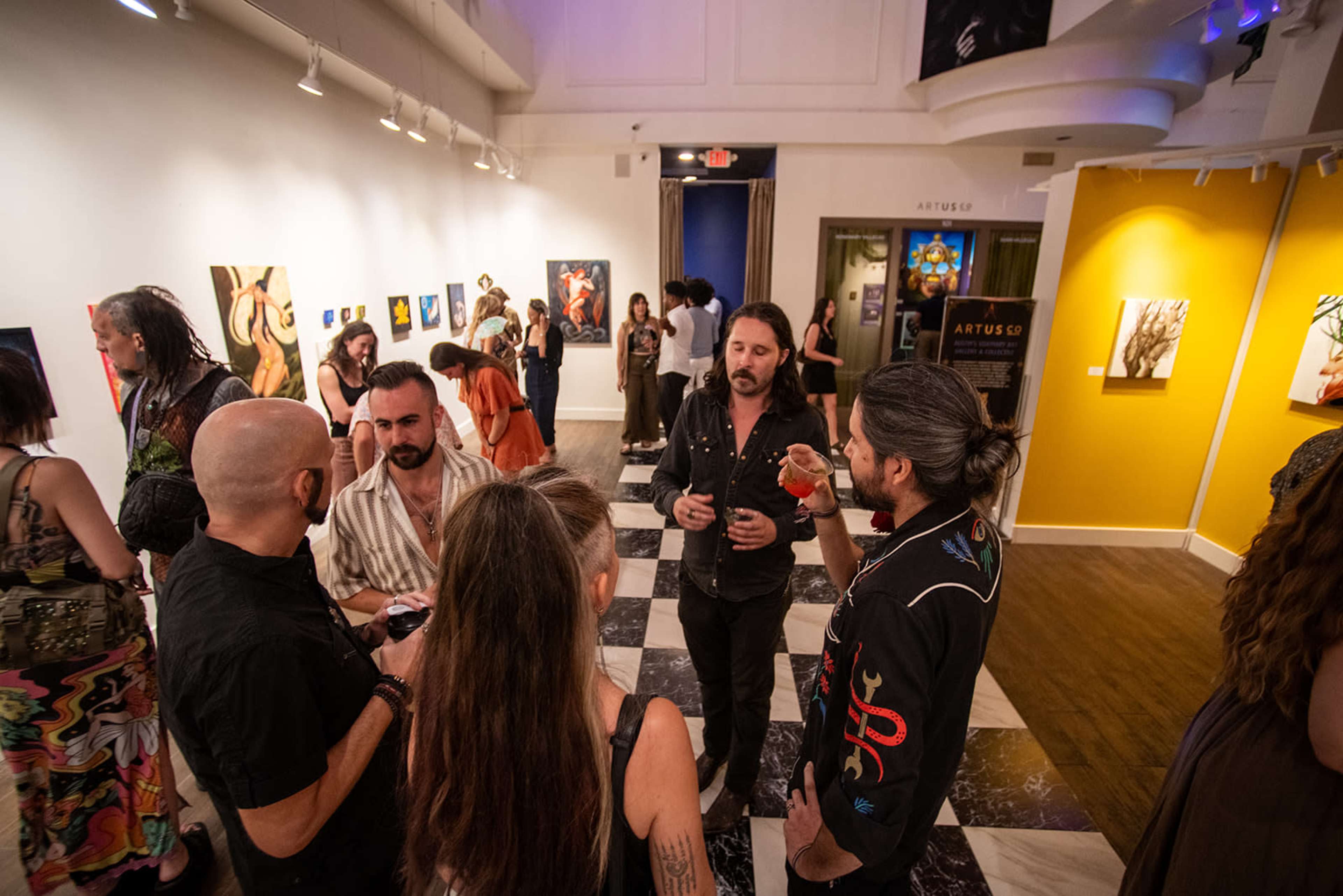 A group of people socialize and network at an art gallery exhibition, surrounded by colorful artworks on the walls.